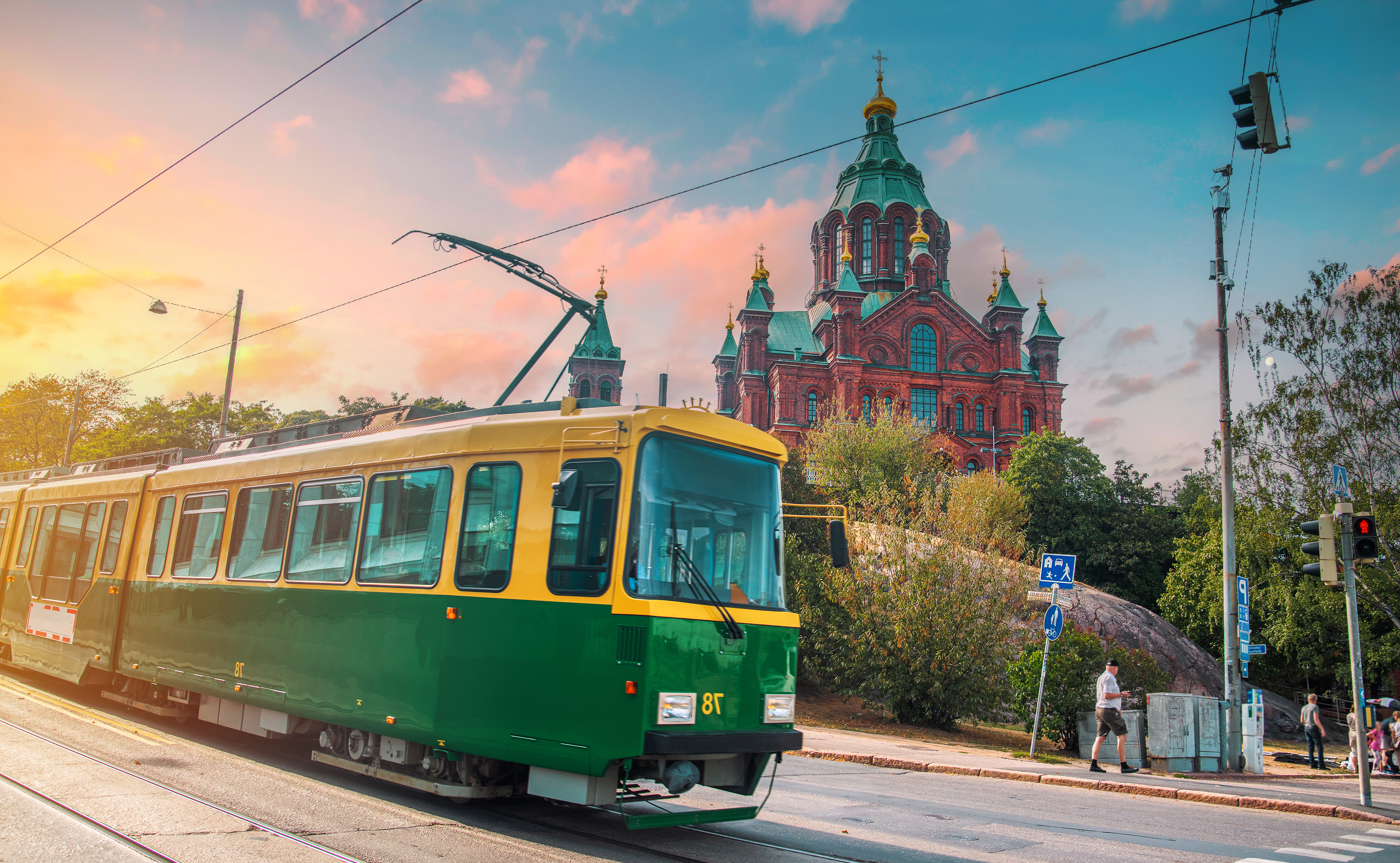 A green and yellow tram travels on a street with a historic building in the background at sunset.