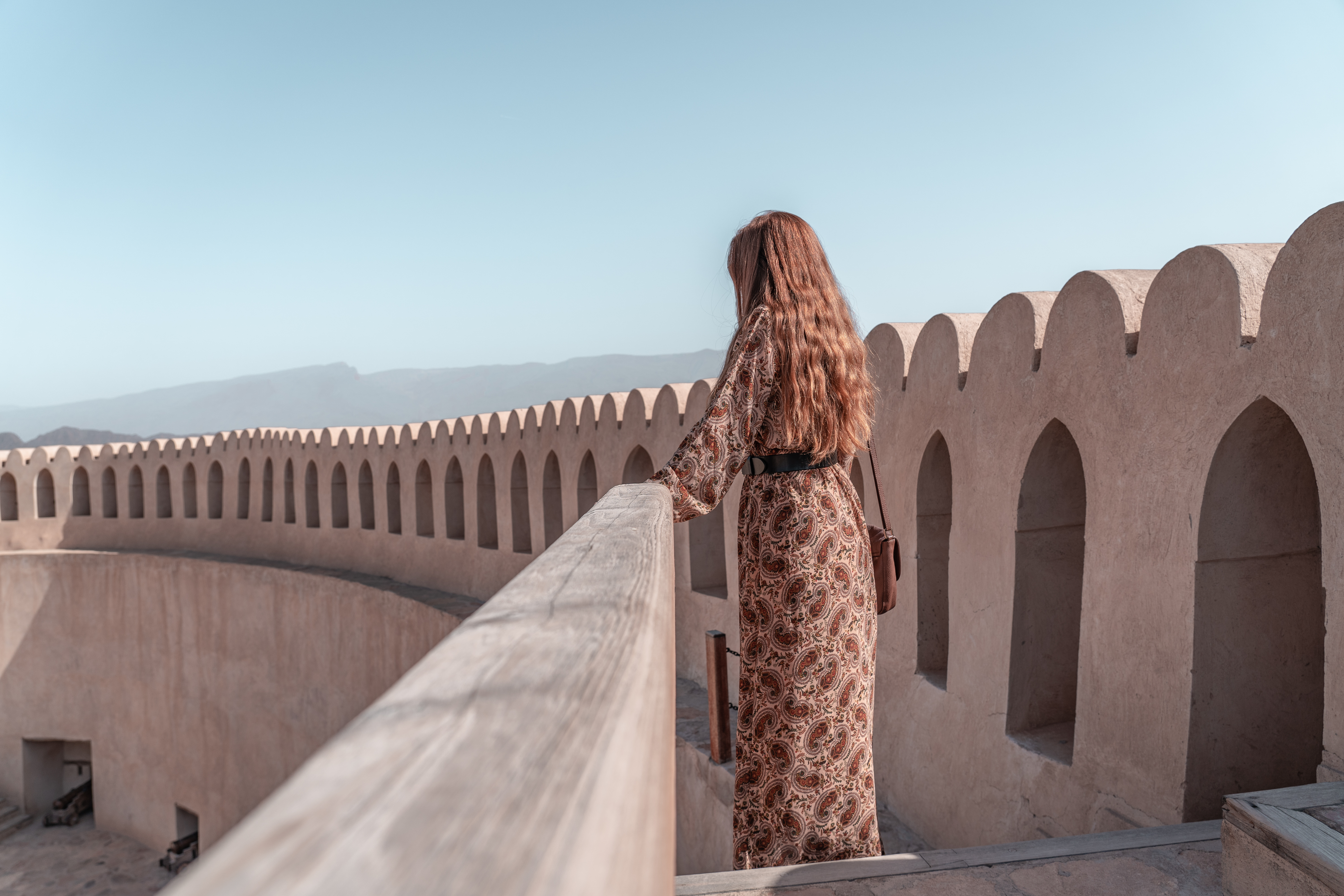 A woman in a patterned dress stands on a balcony, gazing at distant mountains under a clear blue sky.