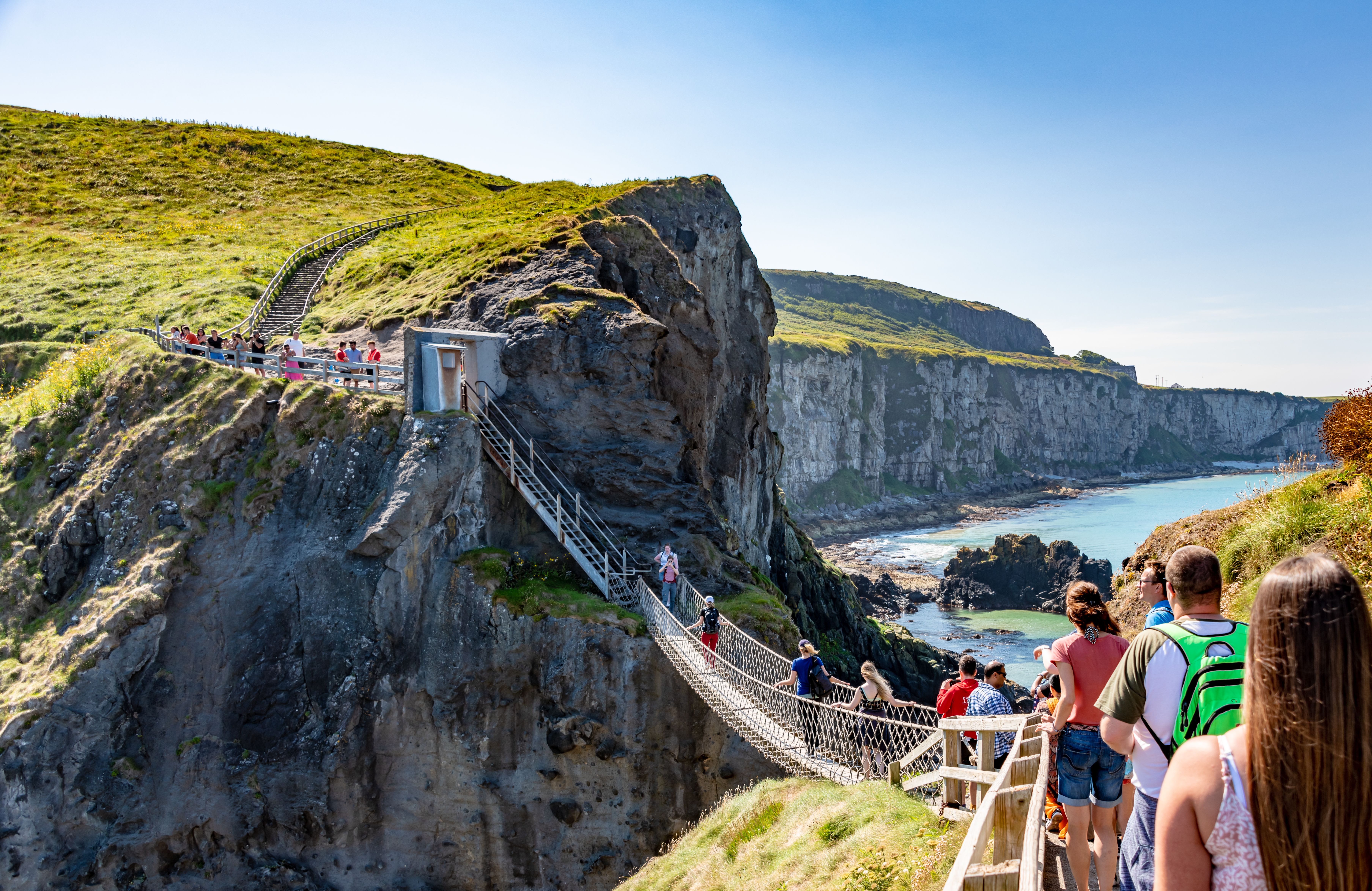 A sunny coastal scene showing a suspended bridge with people walking towards scenic cliffs and the ocean in the background.