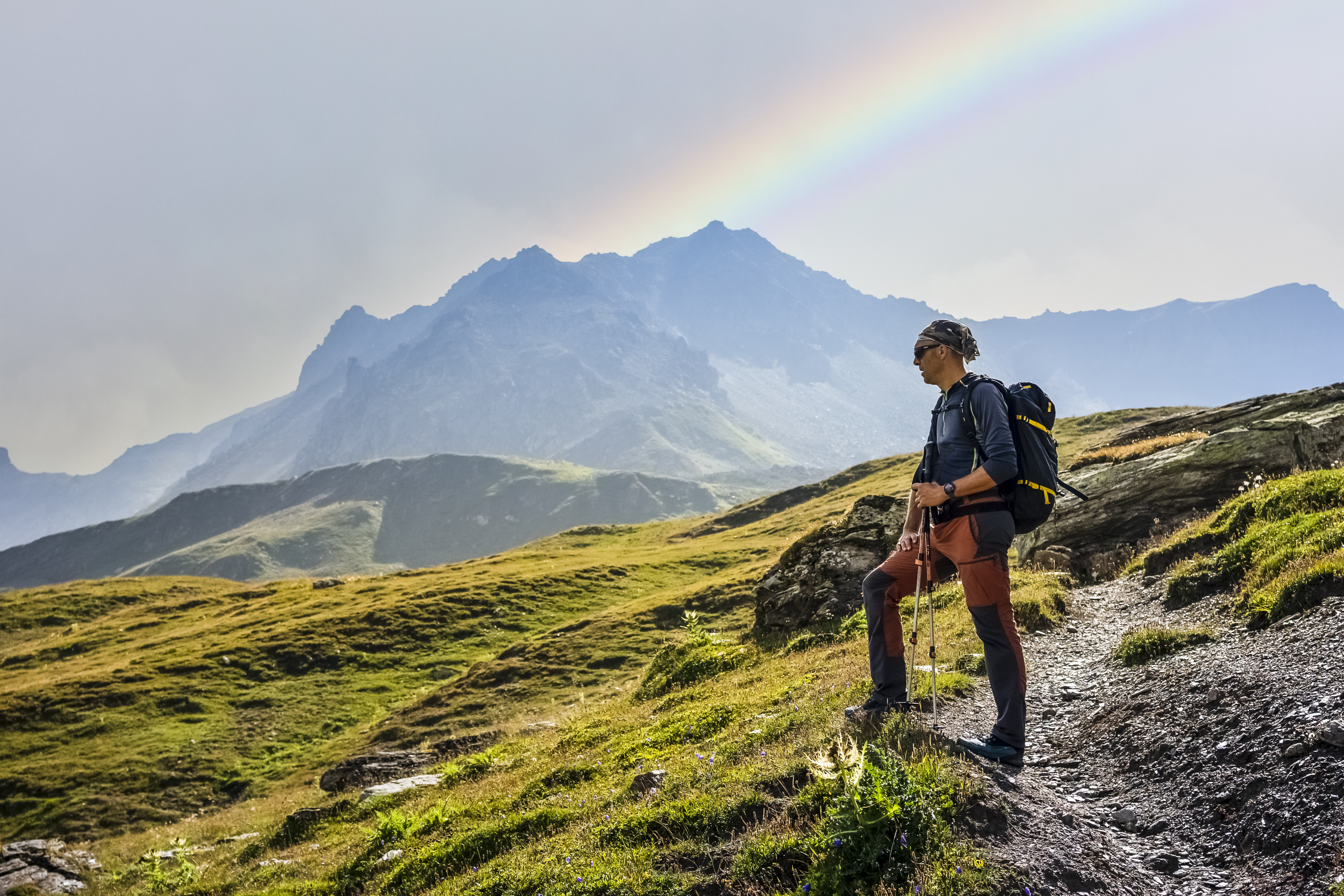 Hiker standing on a rocky trail, gazing at mountains and a rainbow in the sky.