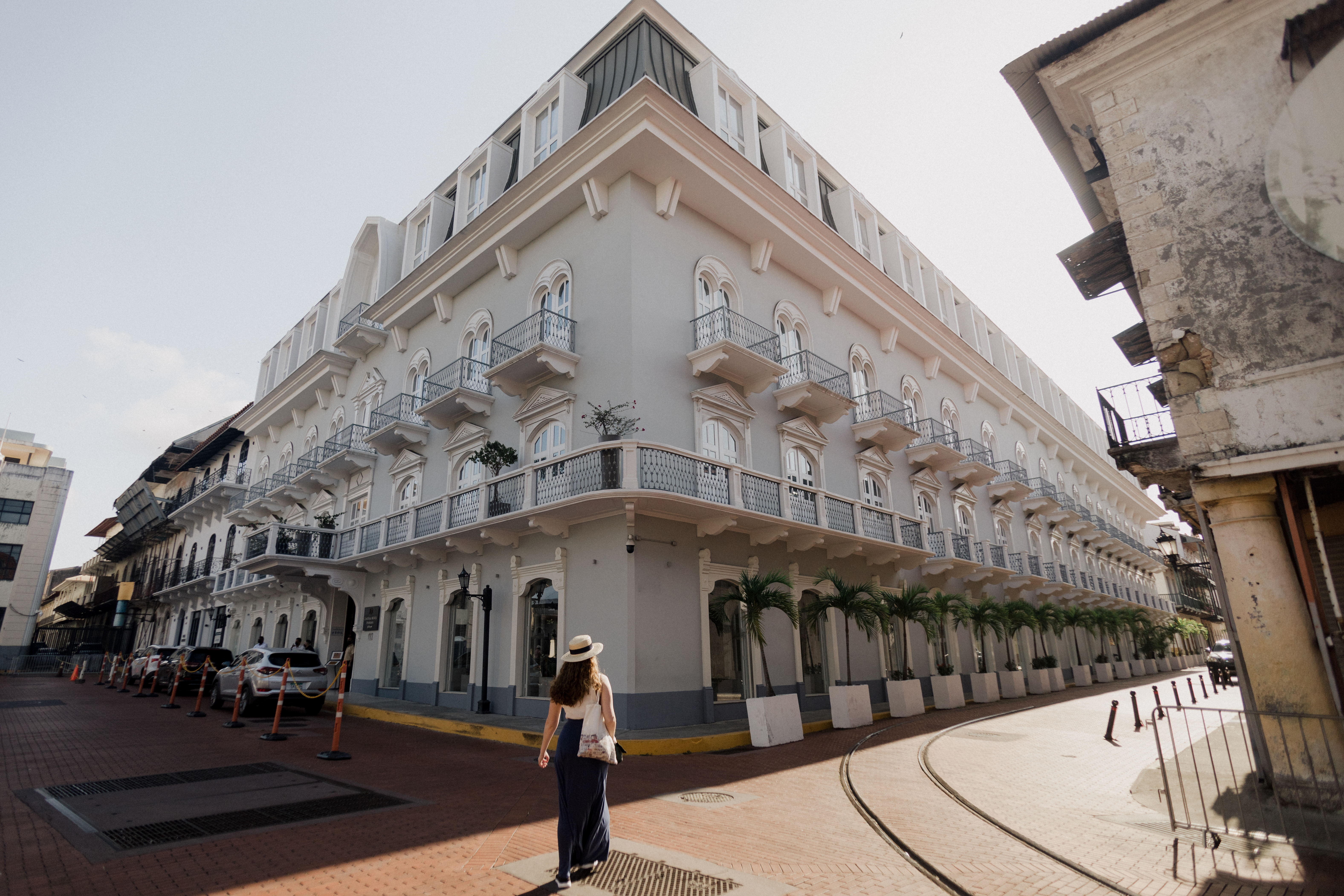 A woman walks along a cobblestone street beside a large, elegant white building with balconies and palm trees.