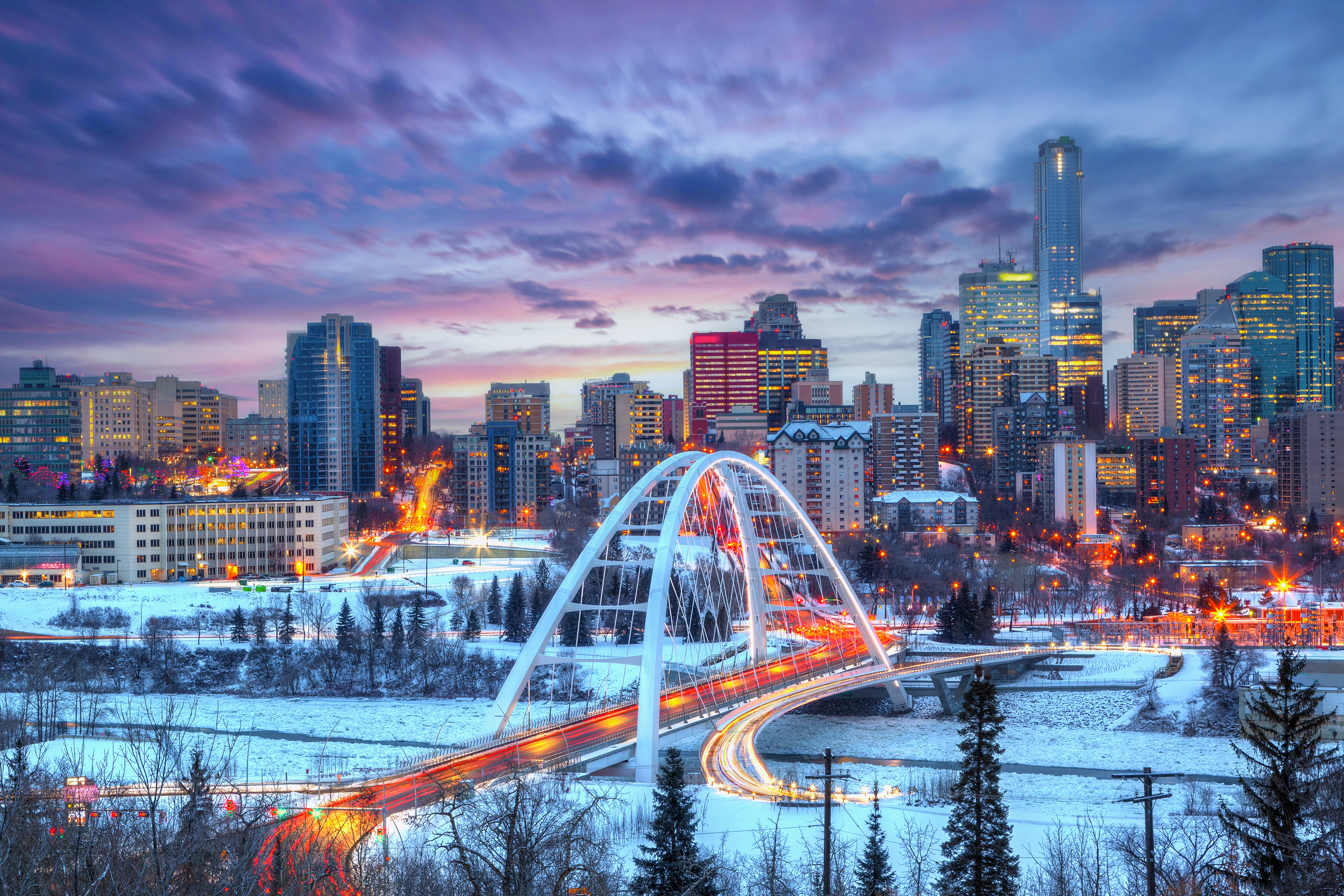 Light trails from rush hour traffic light up downtown Winter sunset skyline showing Walterdale Bridge across the frozen, snow-covered Saskatchewan River and surrounding skyscrapers.
