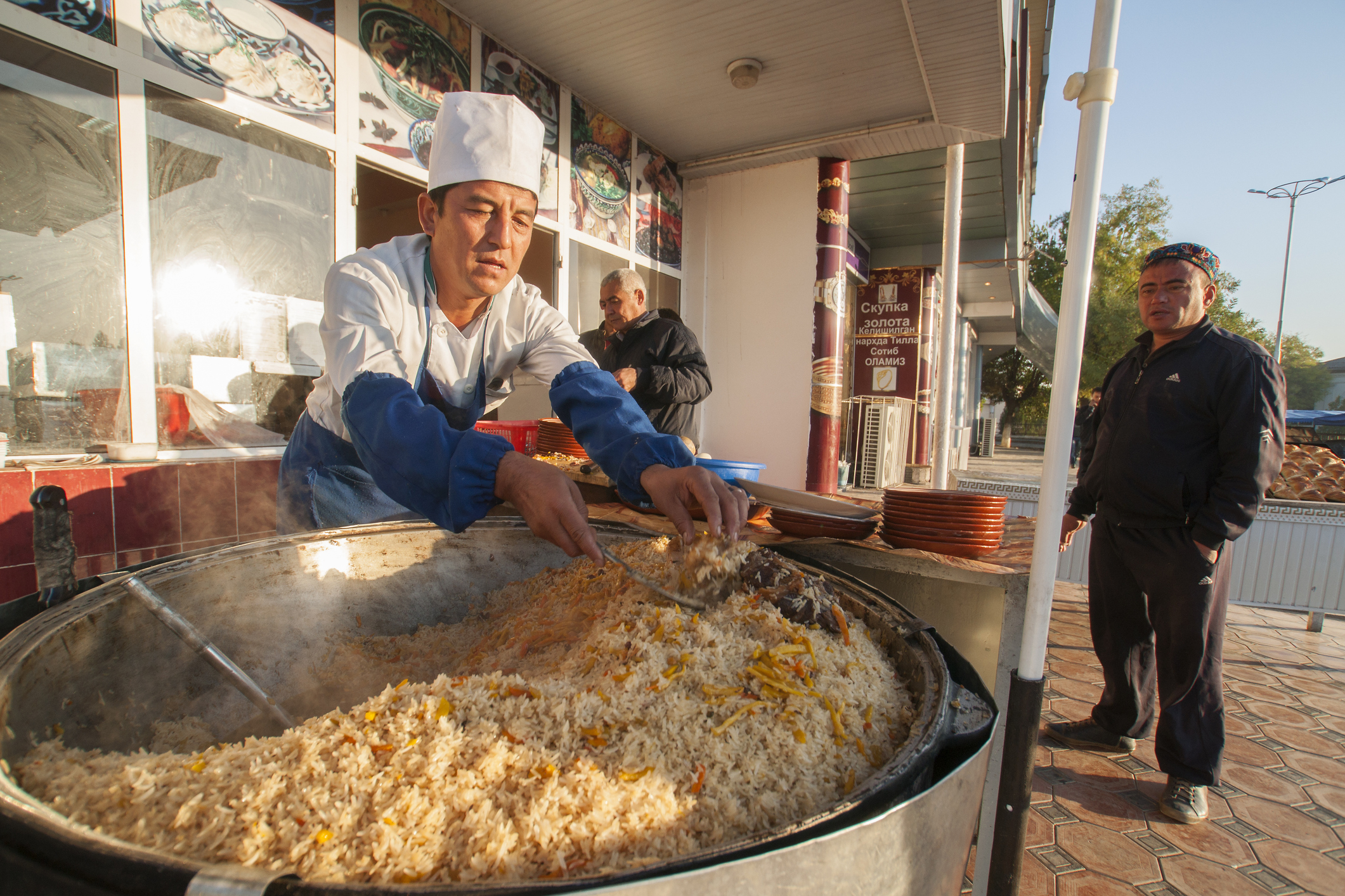 A chef in a white hat prepares a large pot of rice dish outdoors, while a man waits nearby.
