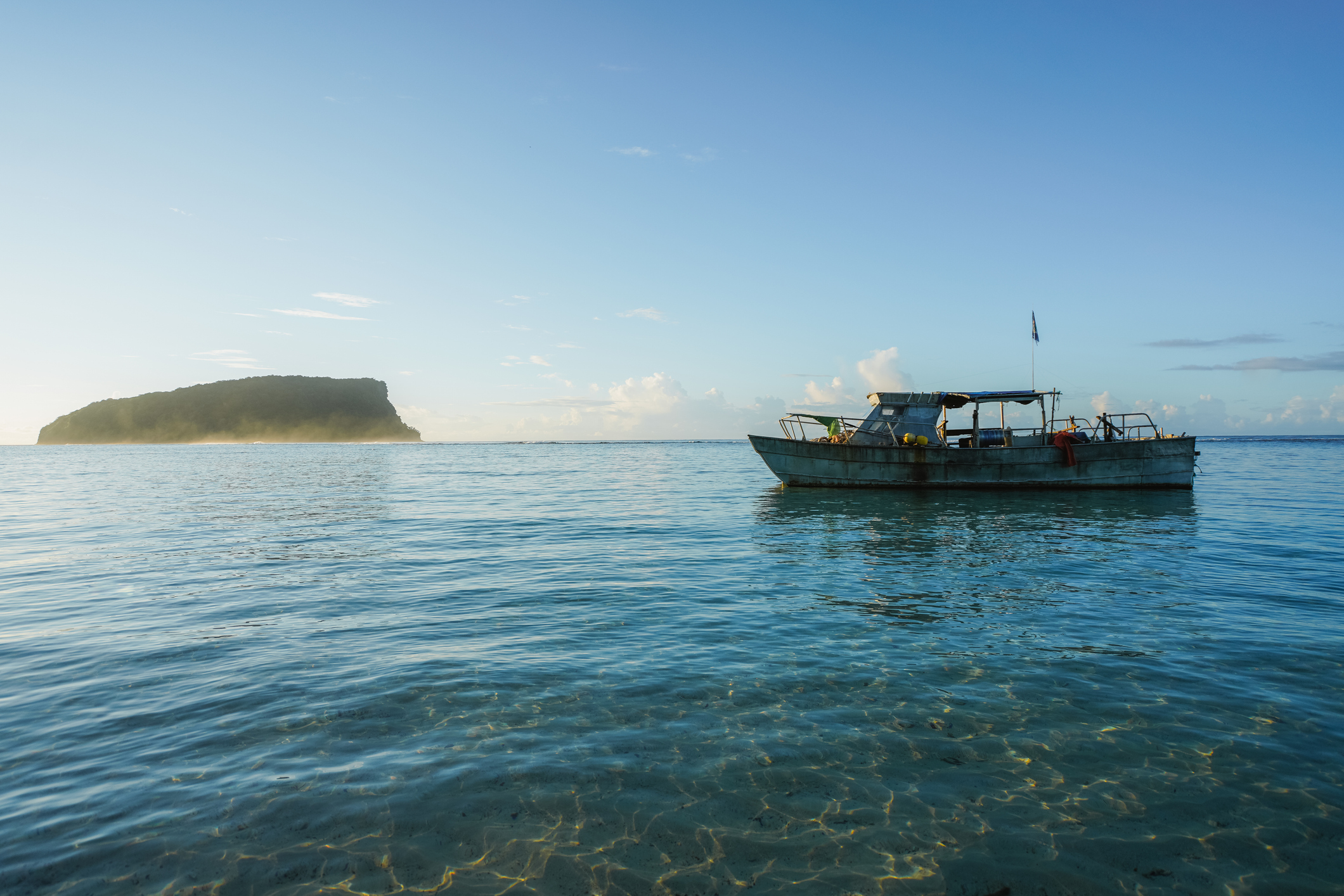 A small boat floats in clear water, with a distant island visible under a bright blue sky.