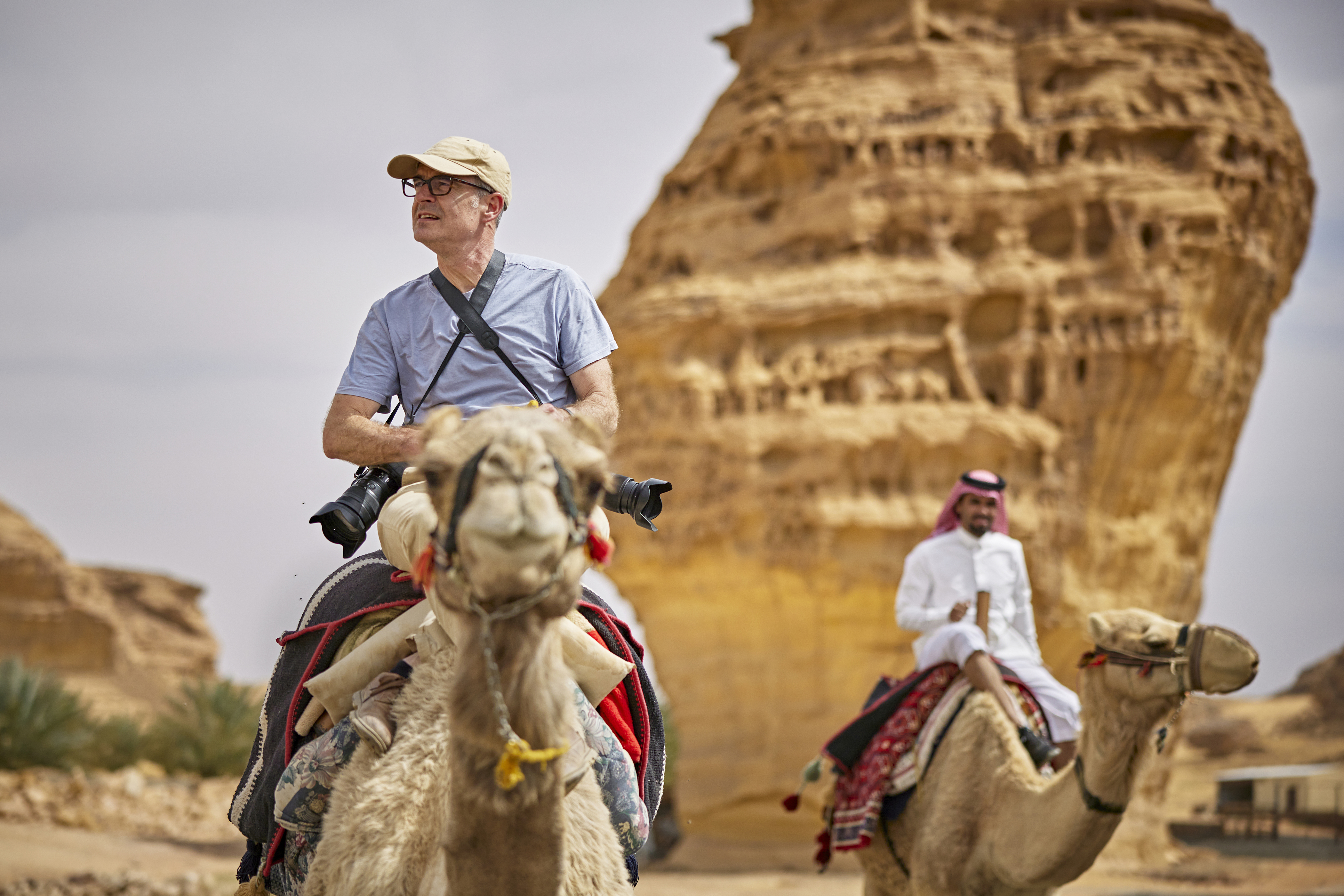 Two men ride camels in a desert landscape, with a large rock formation in the background. One man wears glasses and a cap.