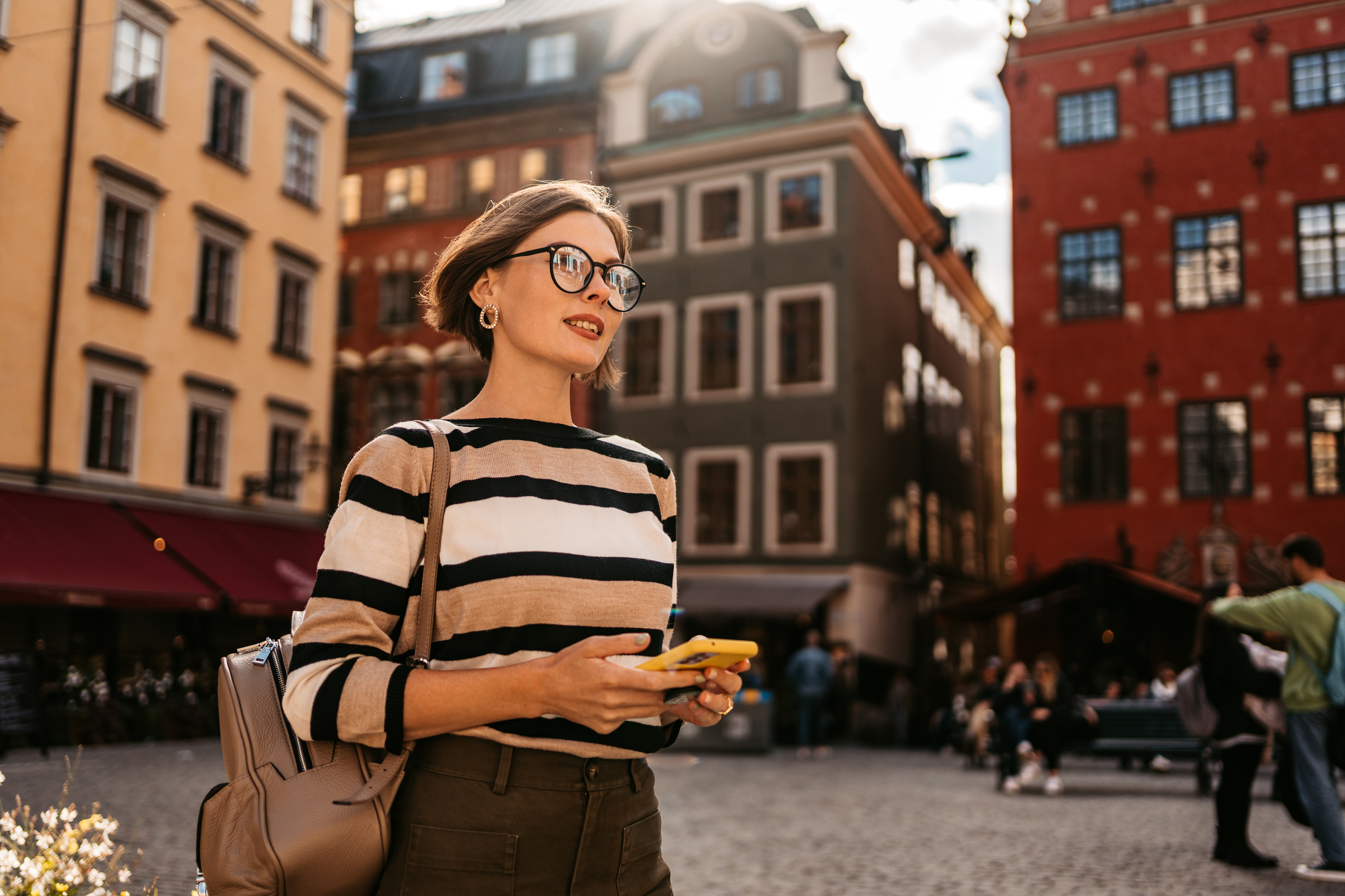 Young woman in a striped sweater stands in a plaza, holding a phone, with colorful buildings and people around her.