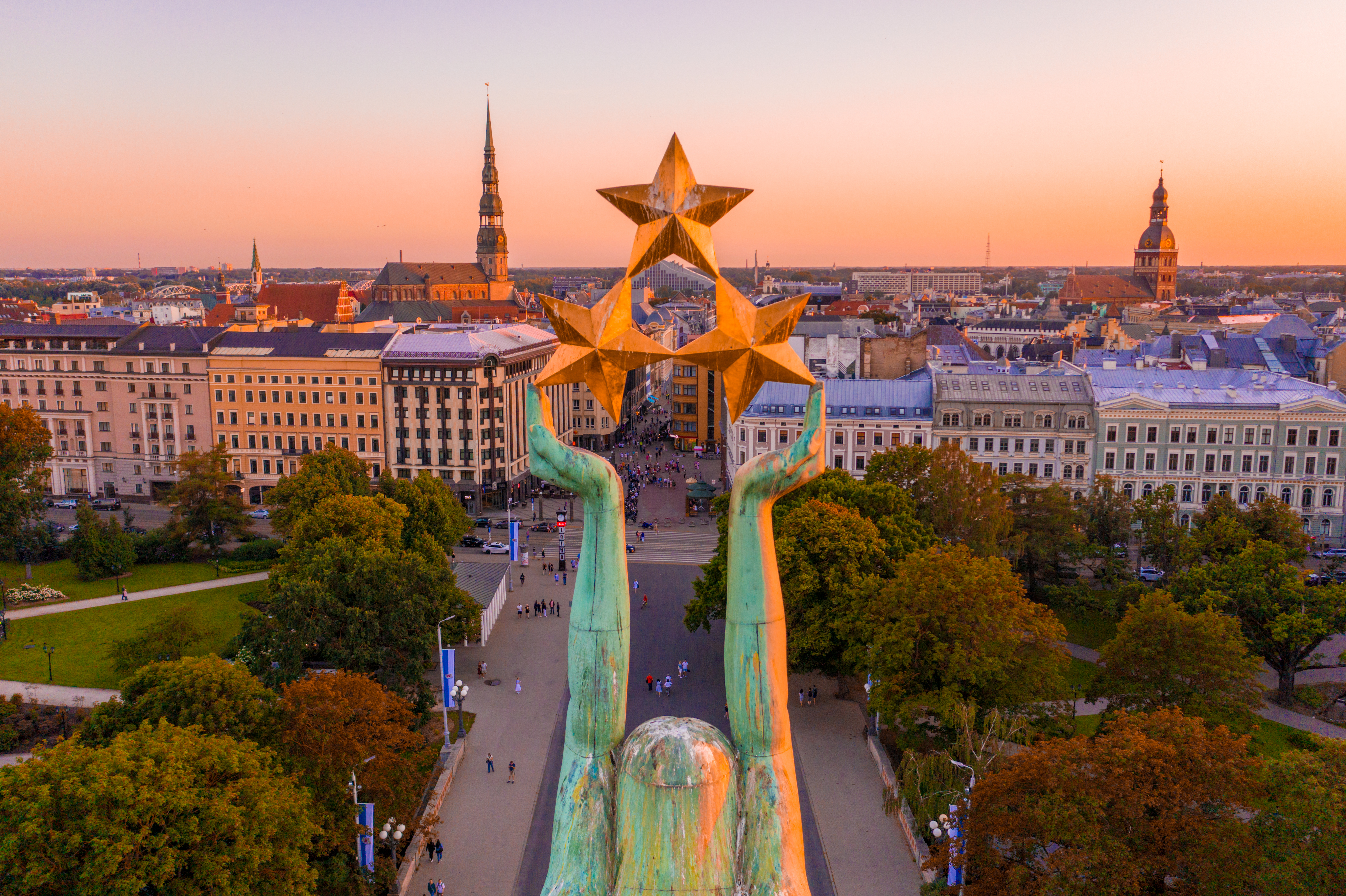 View of a city at sunset, featuring a monumental statue with raised hands holding three stars, surrounded by trees and buildings.
