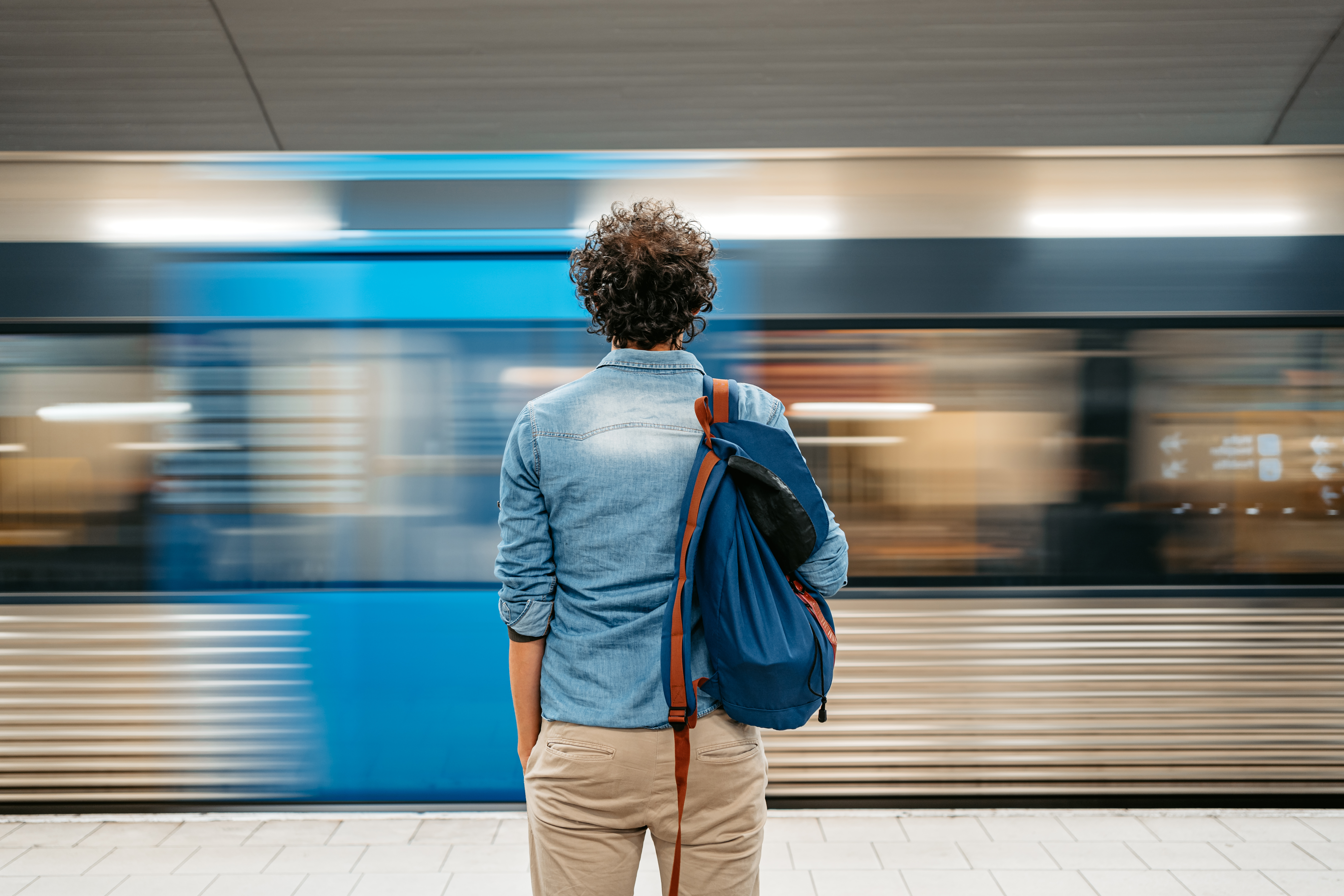 A person stands at a subway platform, facing a train speeding by, with a blue bag slung over their shoulder.