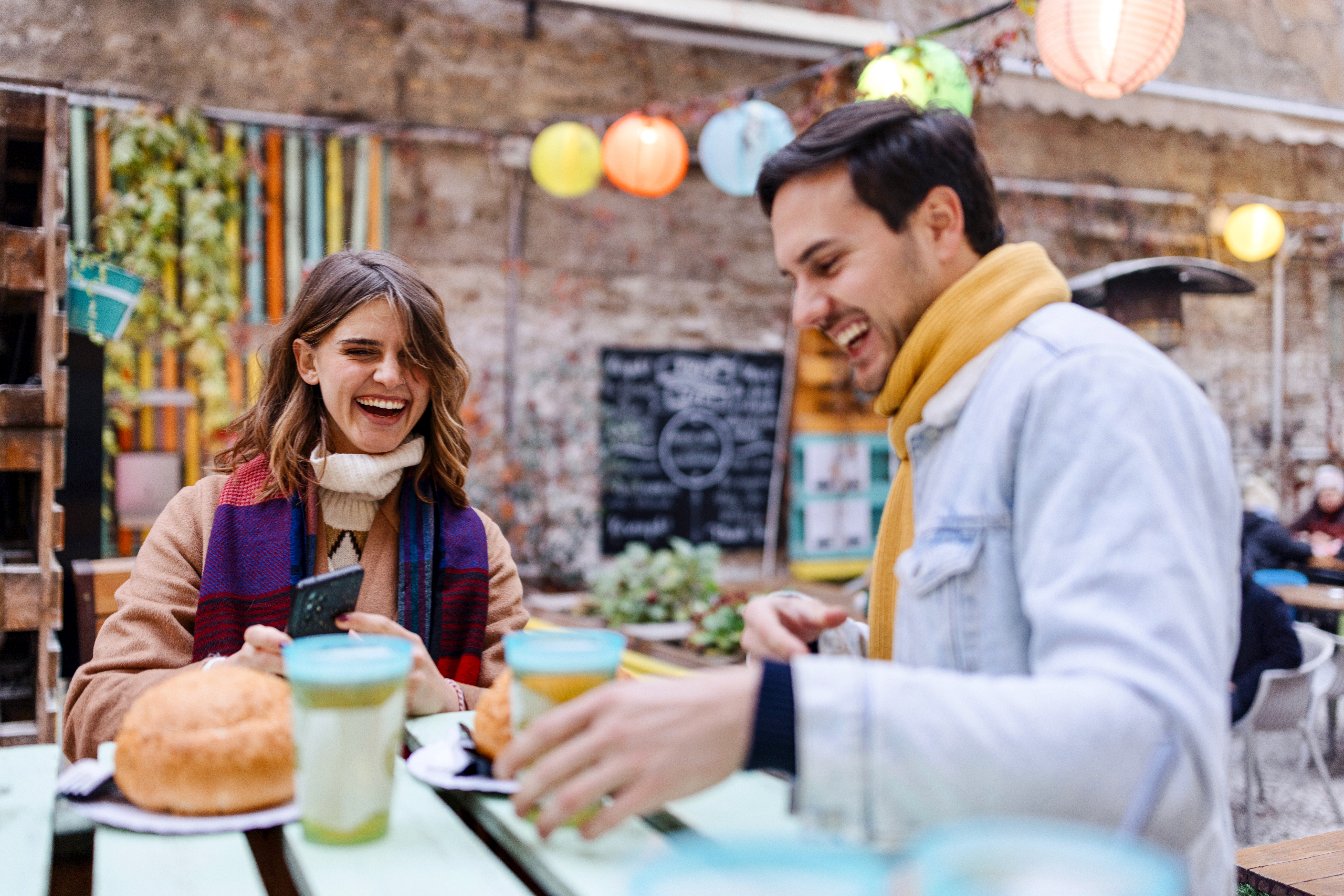 A man and woman sit at a colorful outdoor cafe, laughing and enjoying drinks and pastries.