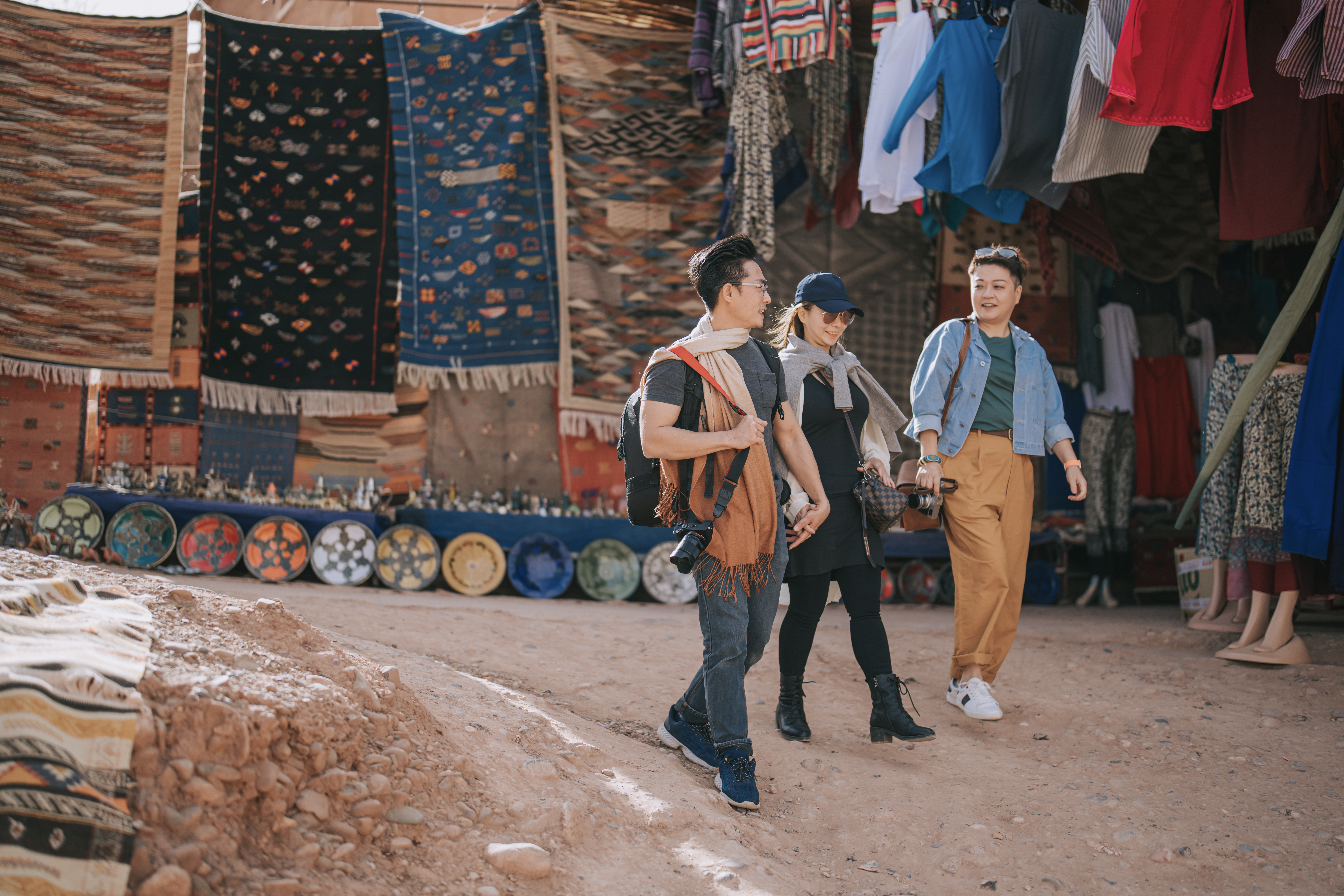 Three people walk through a vibrant market, surrounded by colorful textiles and handmade pottery.