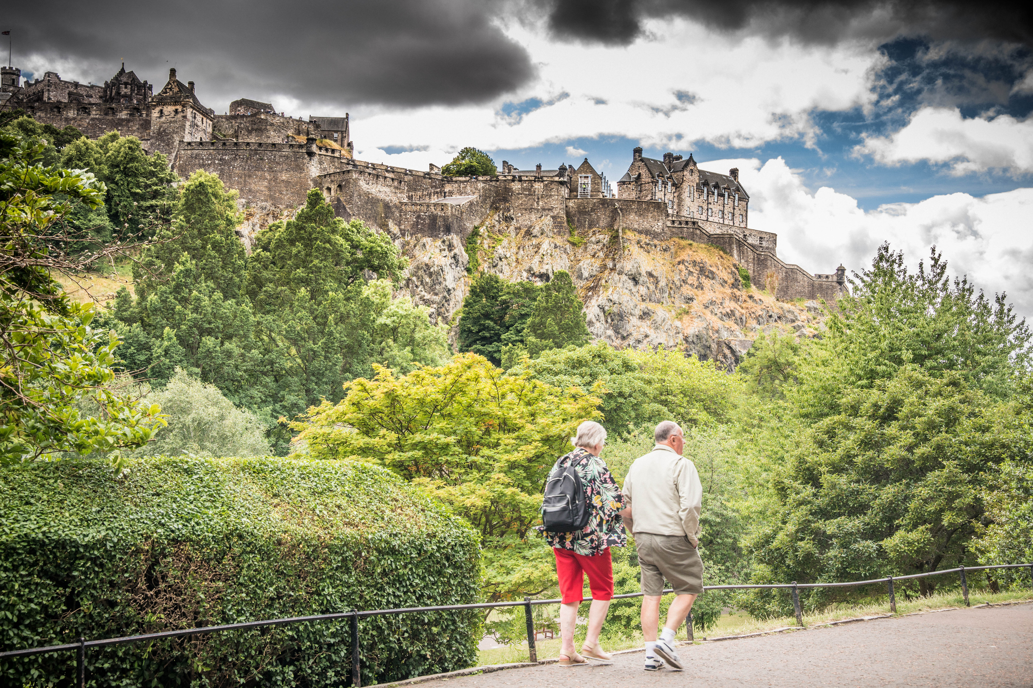 Two people stroll along a path, with Edinburgh Castle perched on a rocky hill in the background, surrounded by greenery.