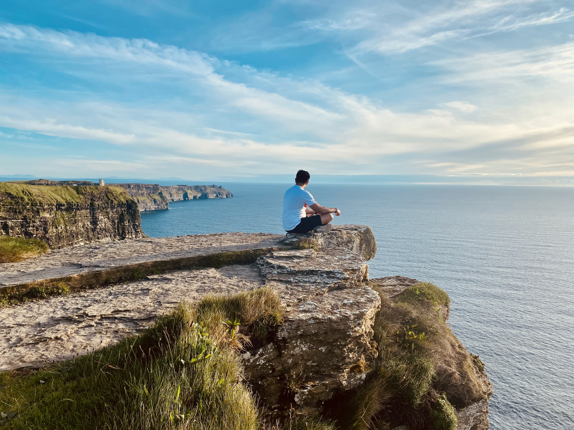 A person sits on a rocky cliff, overlooking a calm sea under a clear sky.