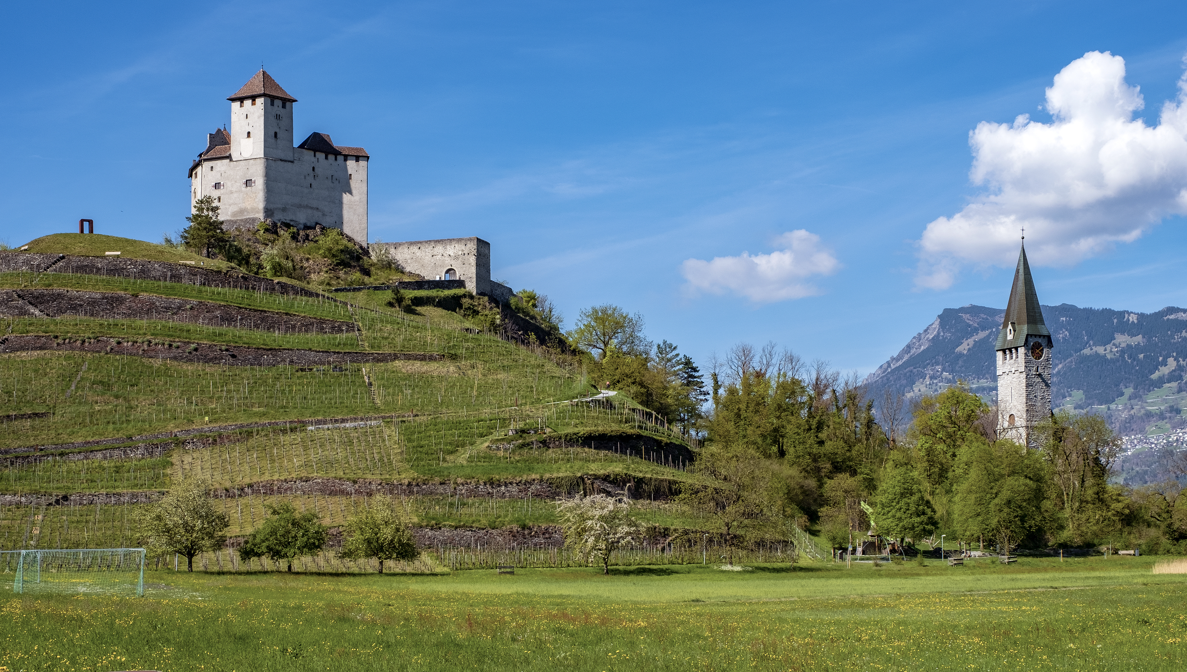A hillside castle and church tower stand against a blue sky, surrounded by greenery and distant mountains.