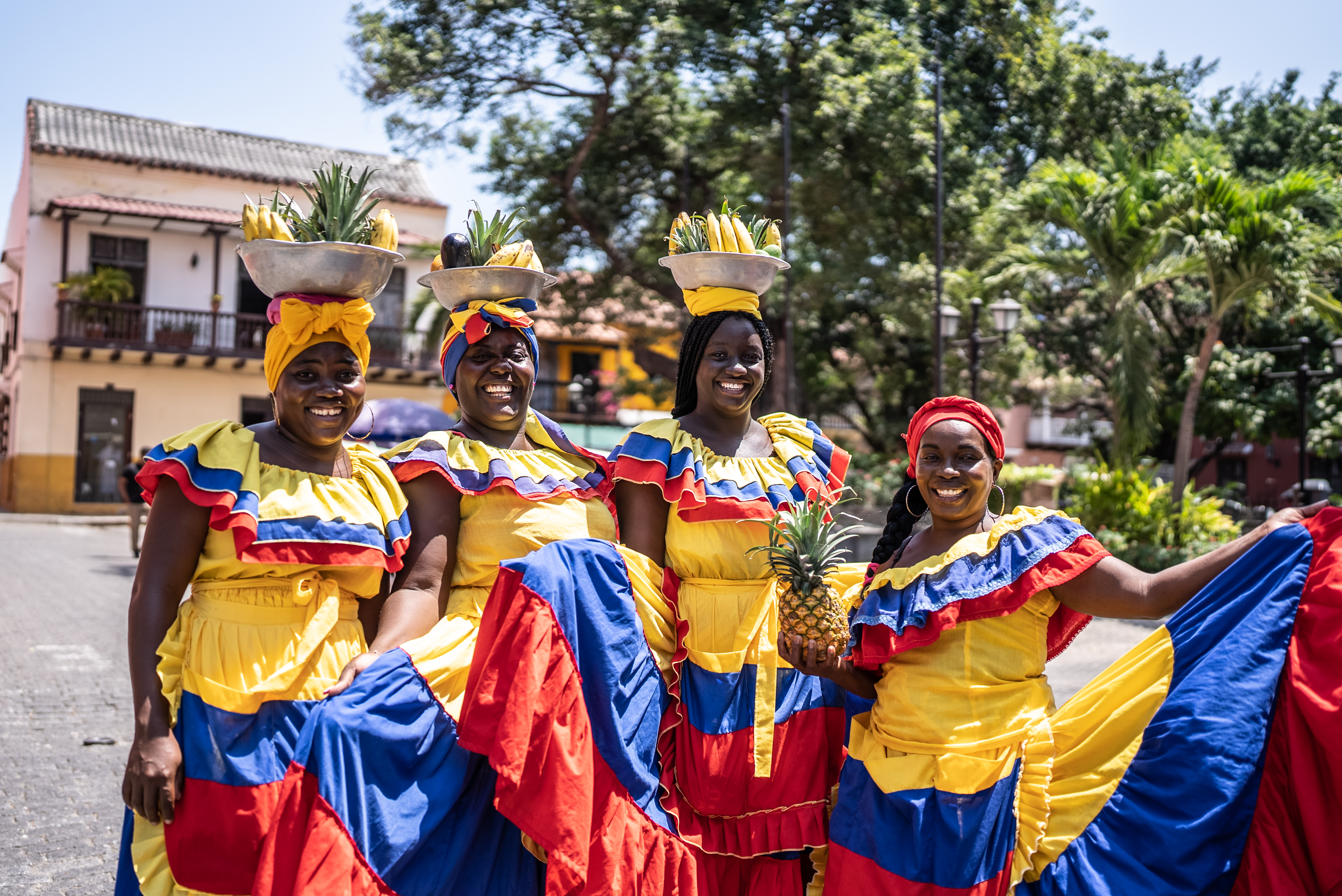 Four women in bright traditional dresses, each carrying a pineapple on their heads, pose happily outdoors.