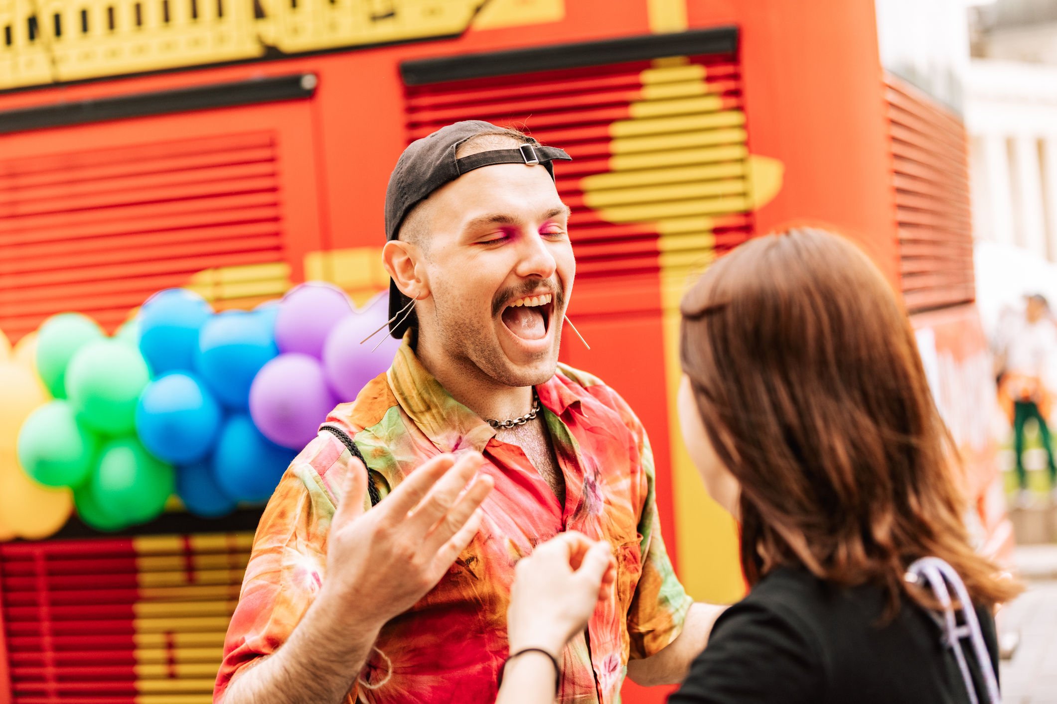 A person with colorful clothing and makeup joyfully interacts with another person against a vibrant, festive backdrop.