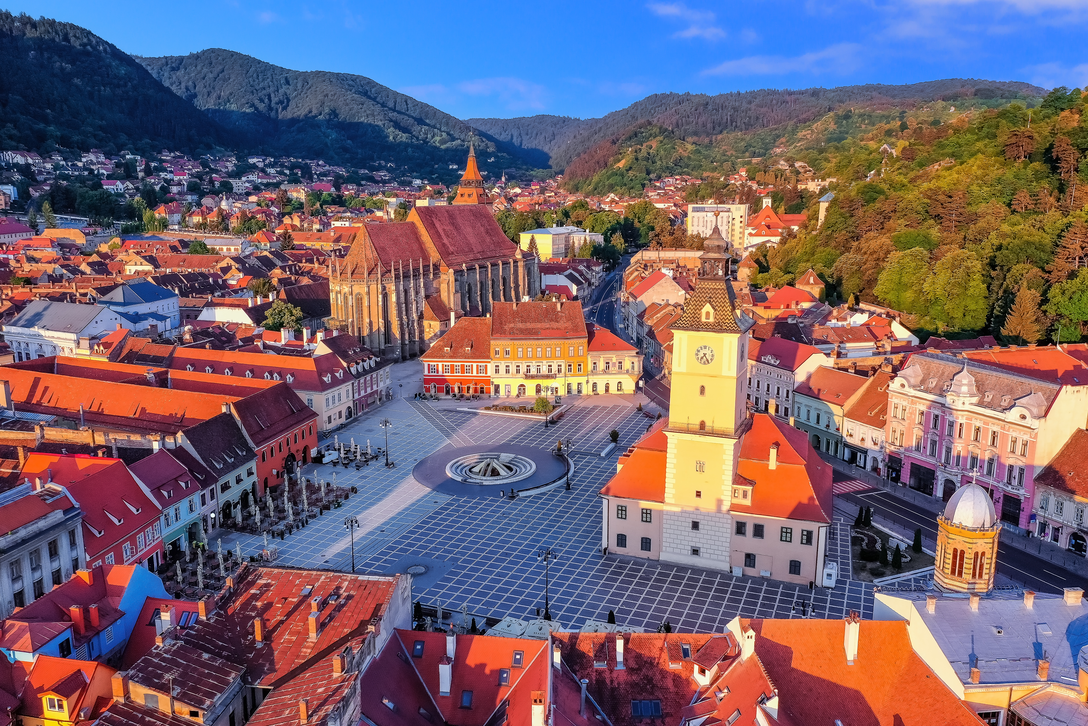 Aerial view of a historic town square with red rooftops, a clock tower, and mountains in the background.