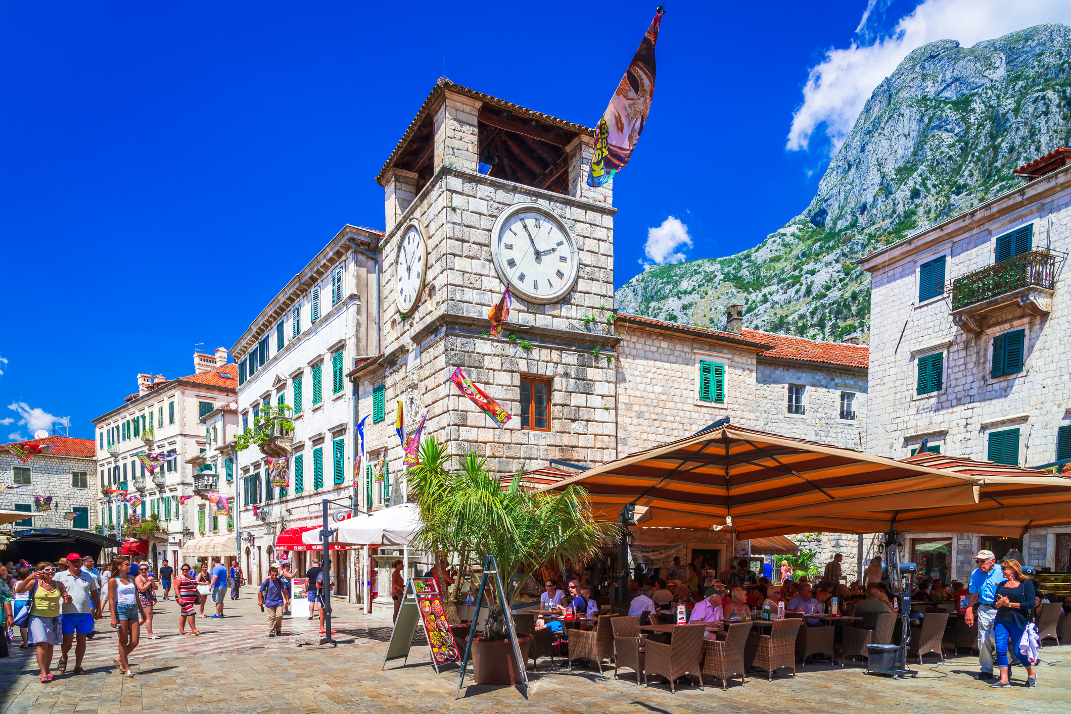 A bustling square in Kotor with a clock tower, cafes, and mountains under a clear blue sky.
