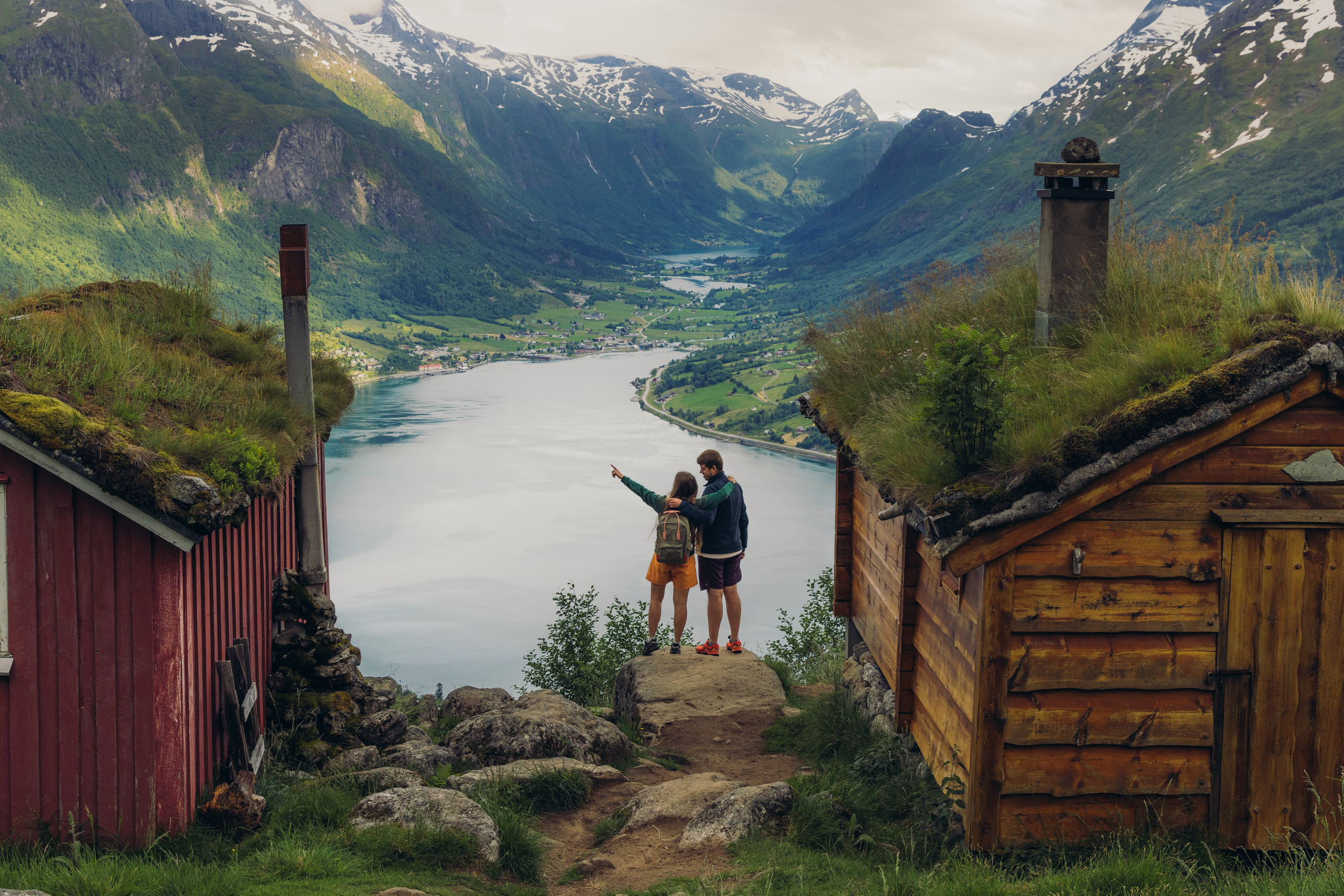 A couple stands between two wooden cabins, overlooking a scenic fjord surrounded by mountains.