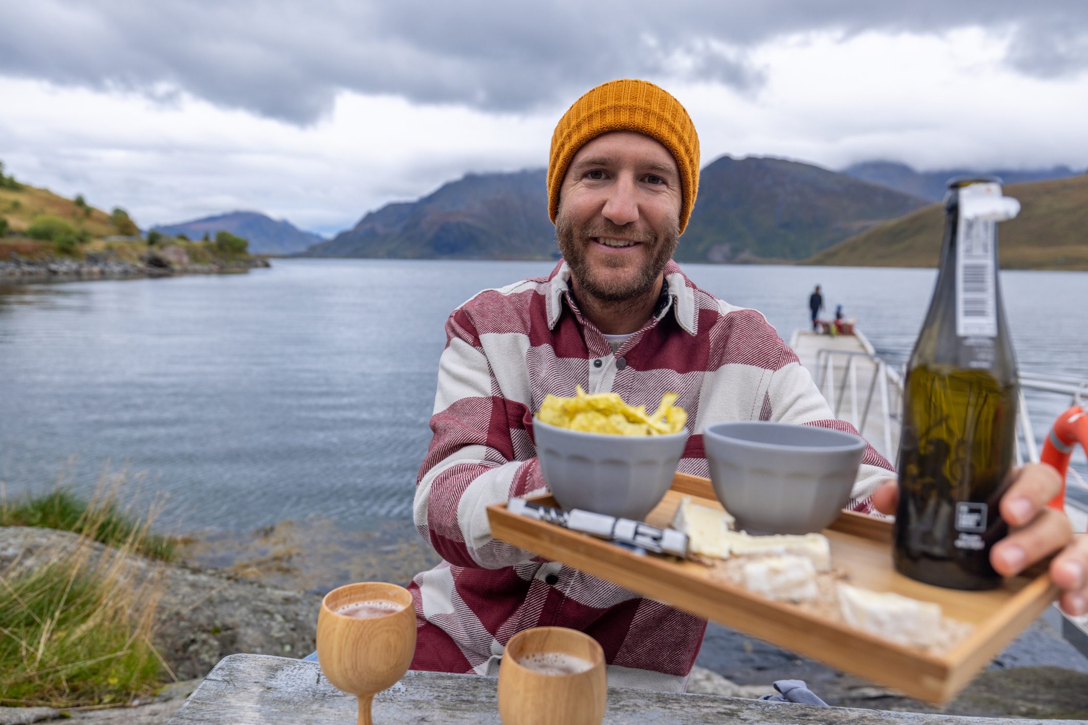 A smiling man in a striped shirt and orange beanie holds a serving tray with food and drinks by a calm lakeside.