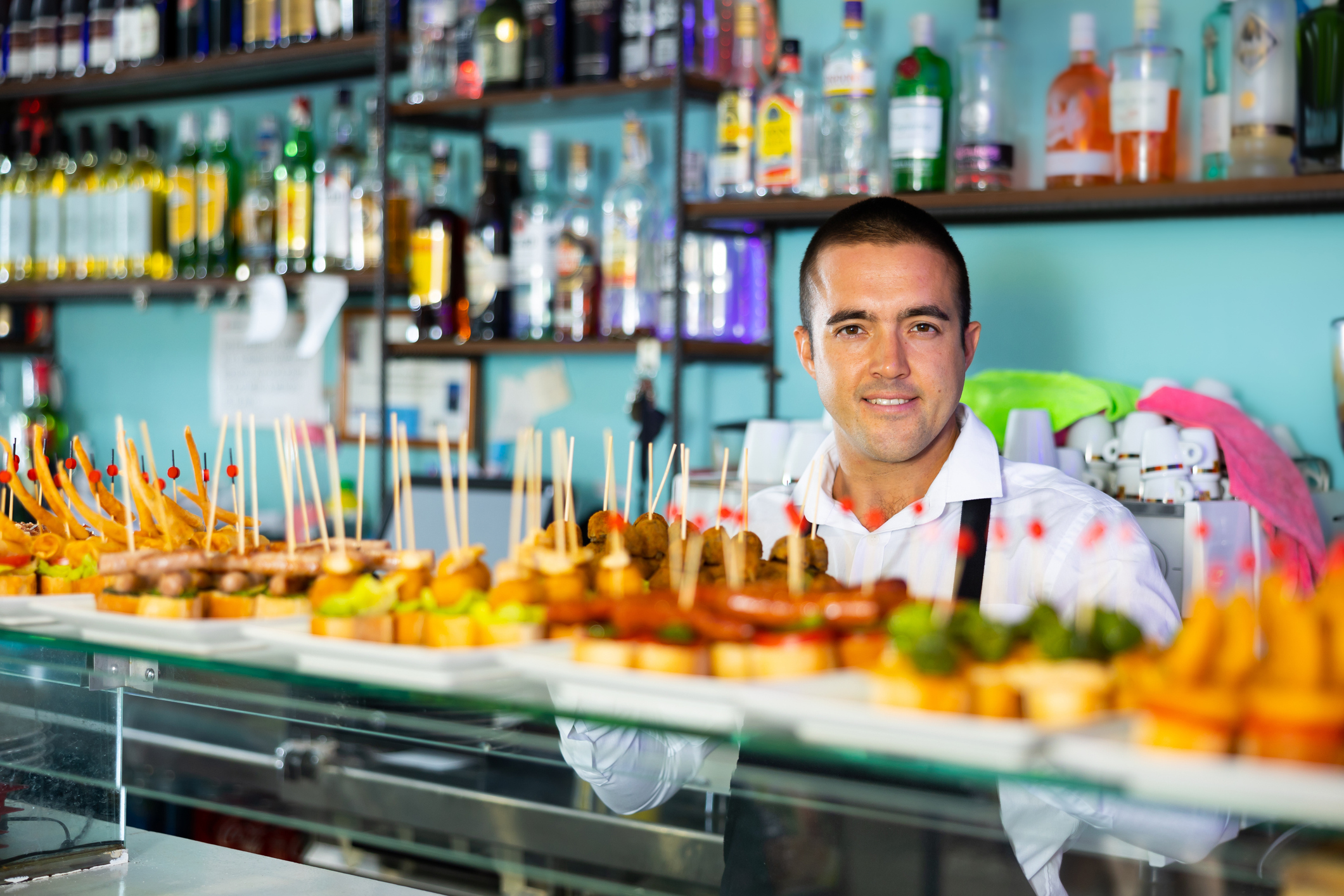 A smiling bartender stands behind a display of colorful tapas skewered with toothpicks, bottles of liquor visible in the background.