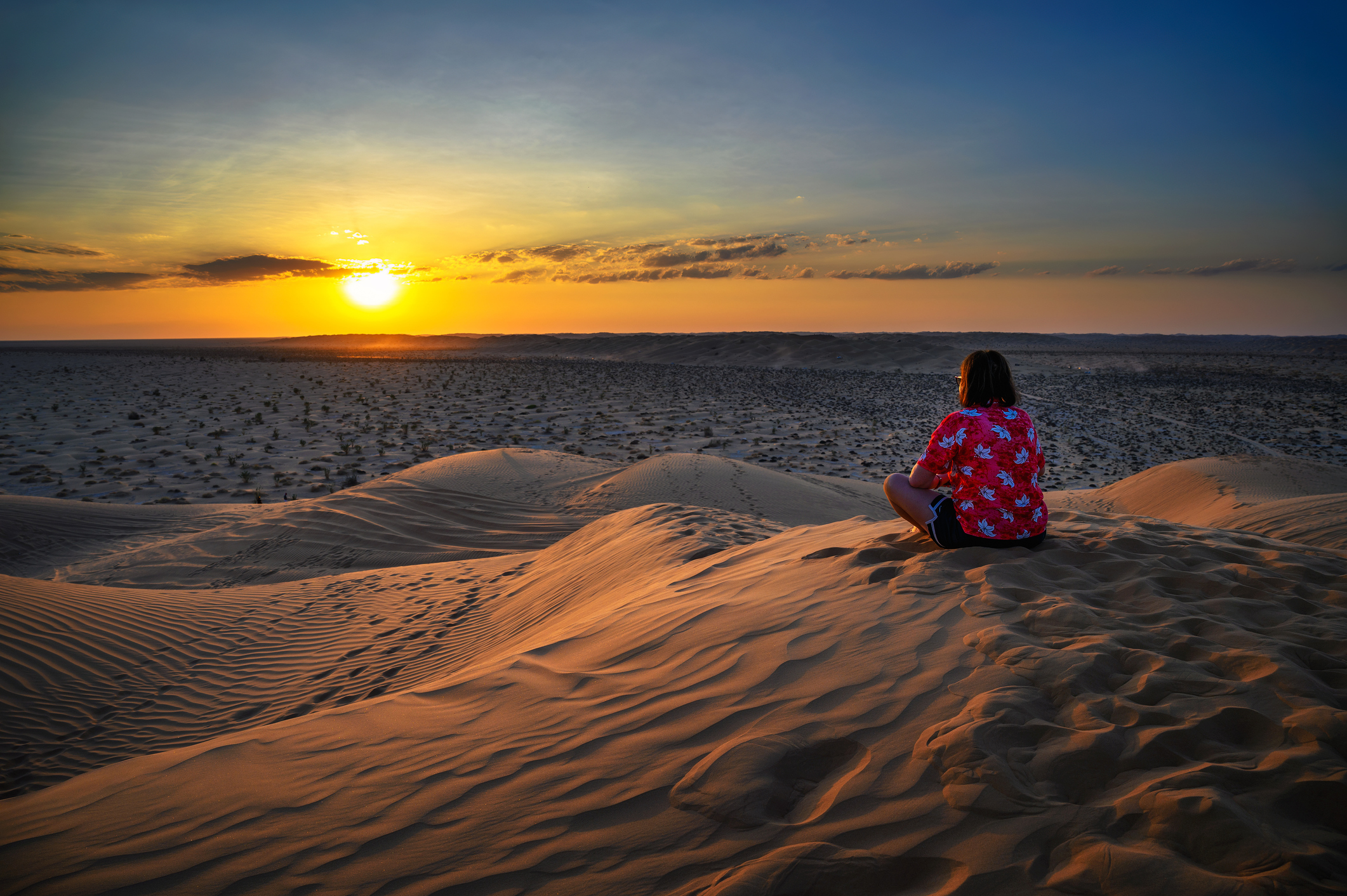 A person sitting on sand dunes, watching a colorful sunset against a vast desert landscape.