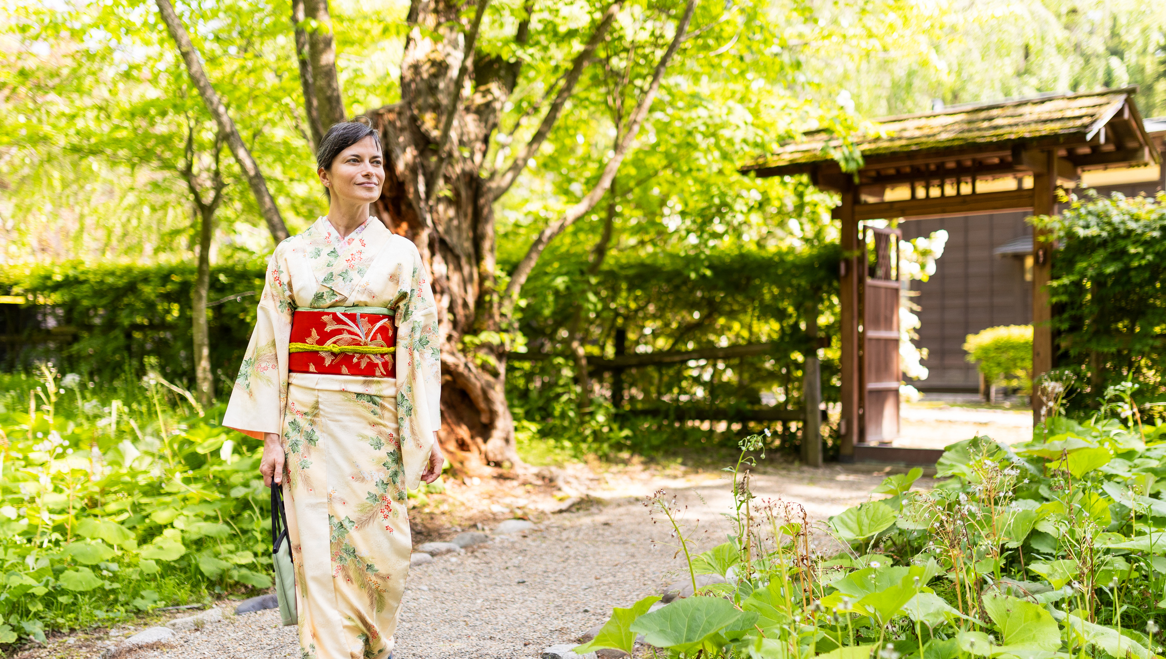 A woman in a traditional kimono walks through a lush, green garden near a wooden gate.