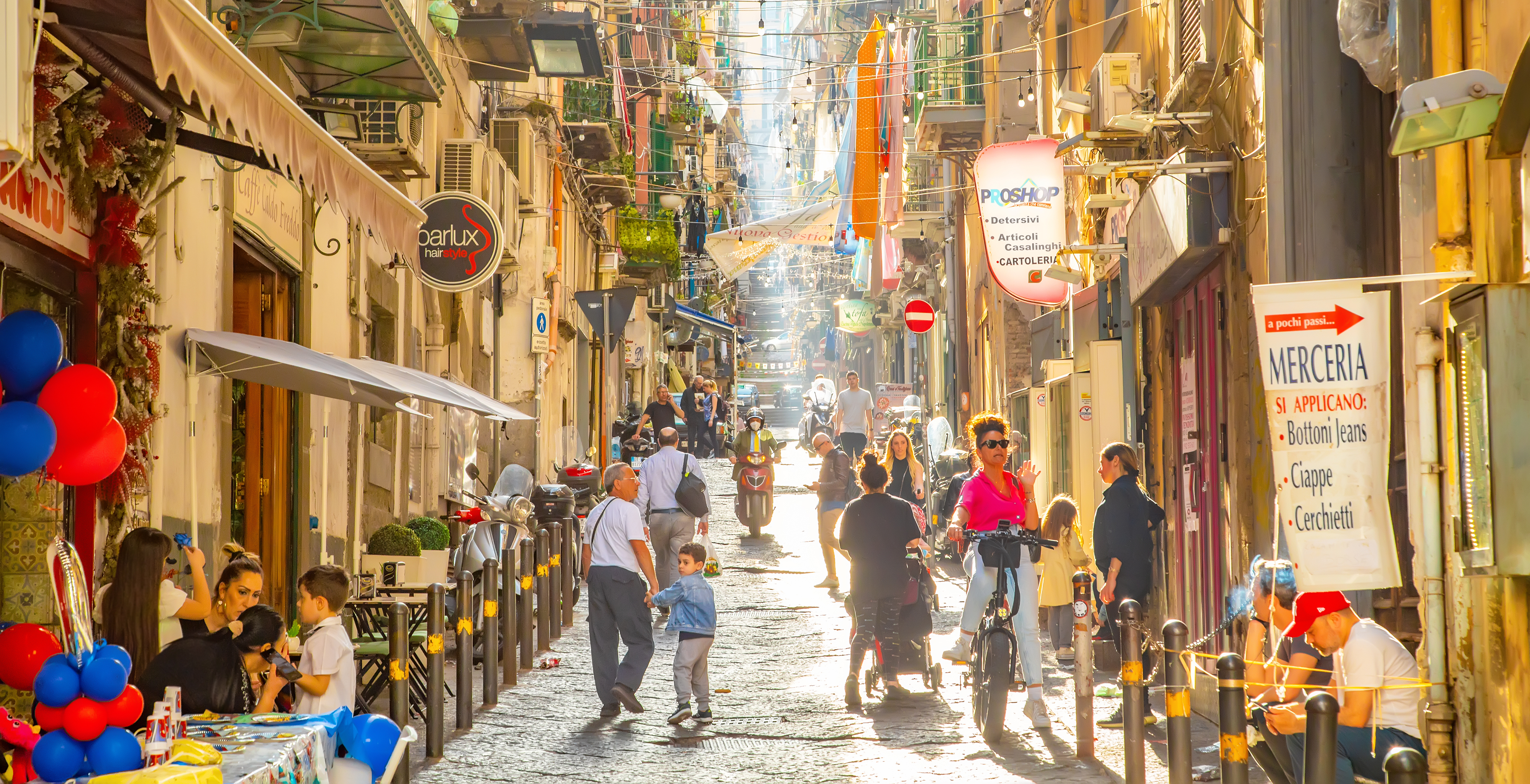 A bustling street scene with people walking, sitting at tables, and colorful decorations under bright sunshine.