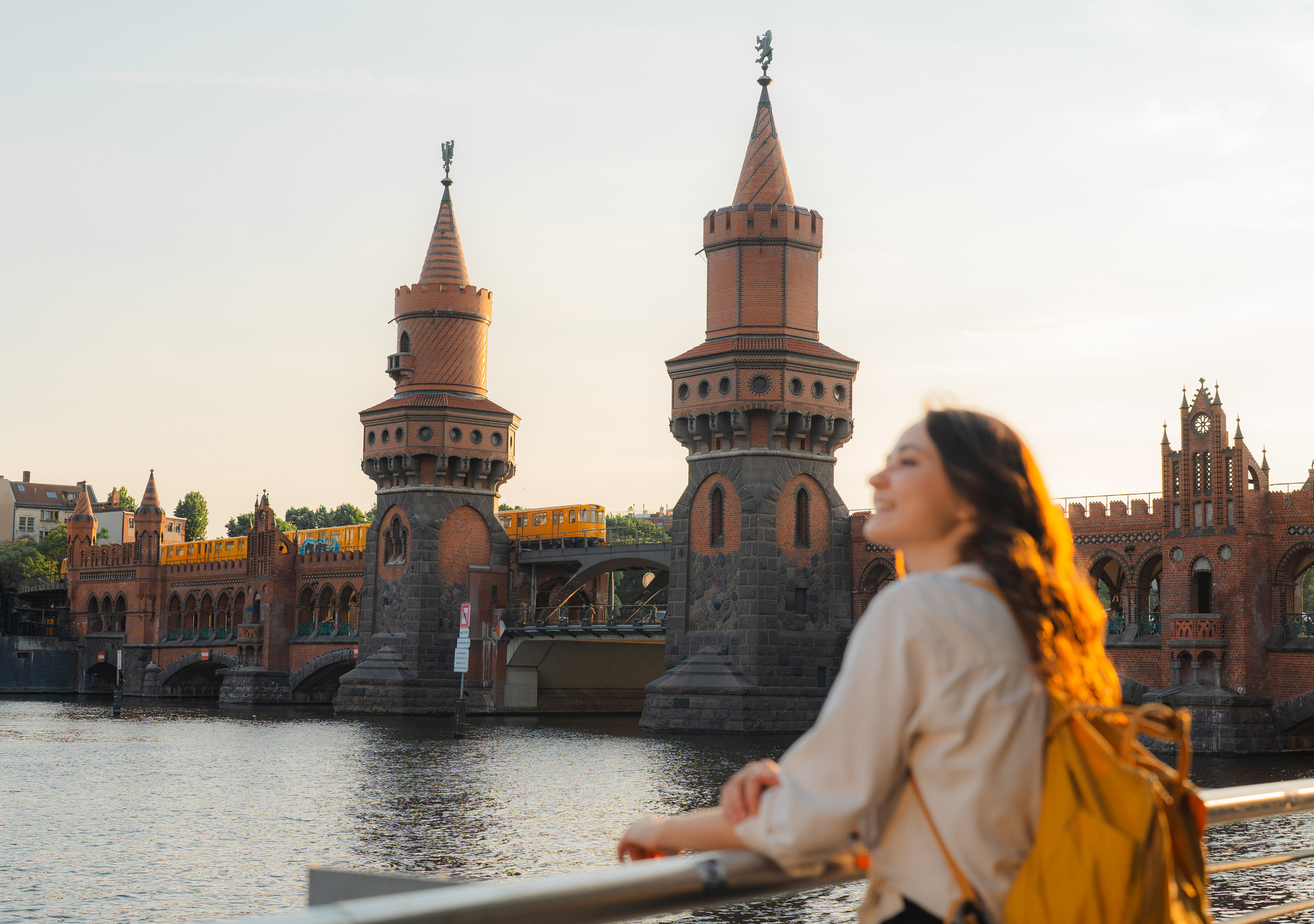 Young woman with a backpack gazes at a historic bridge with towers above a river during sunset.