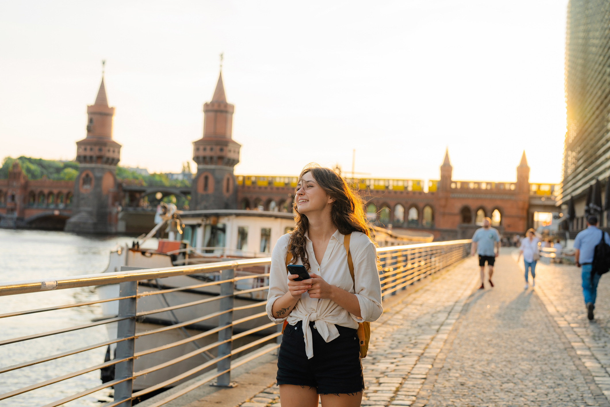 A woman stands on a riverside path, checking her phone, with a bridge and sunset in the background.