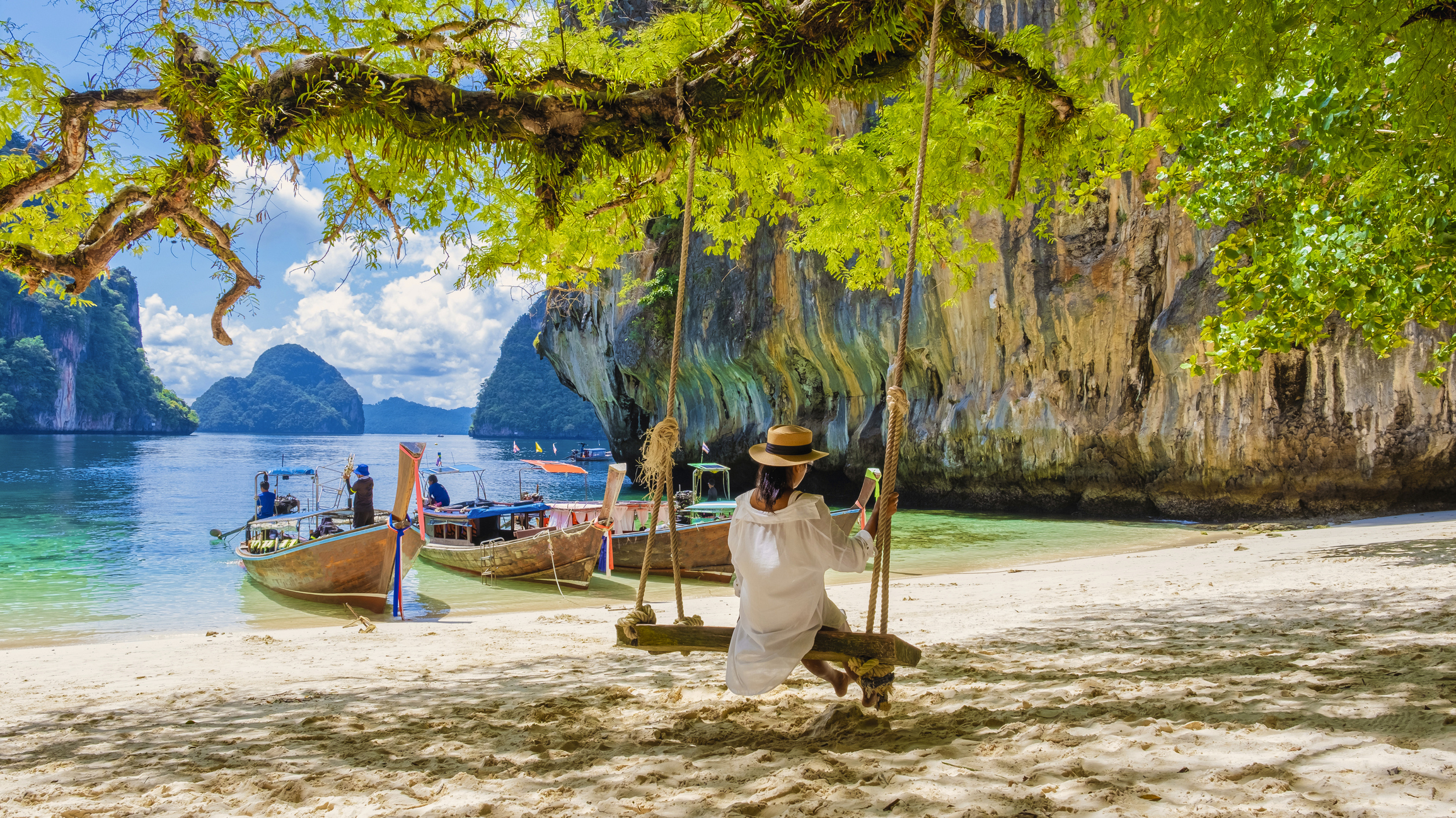 A person on a swing overlooking a tropical beach with boats and limestone cliffs in the background under a sunny sky.