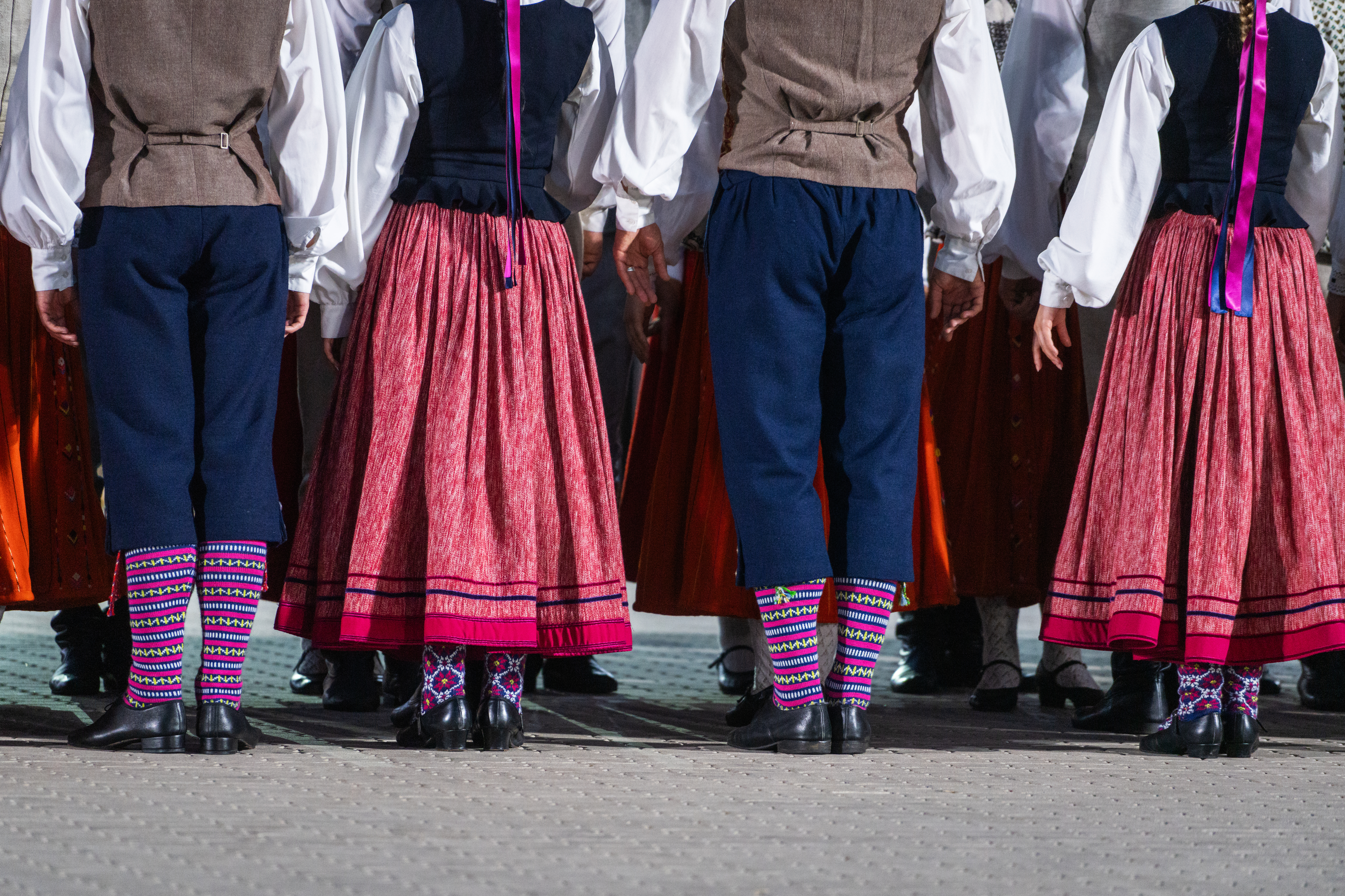 Group of dancers in traditional attire, showcasing colorful skirts and patterned socks from the back.