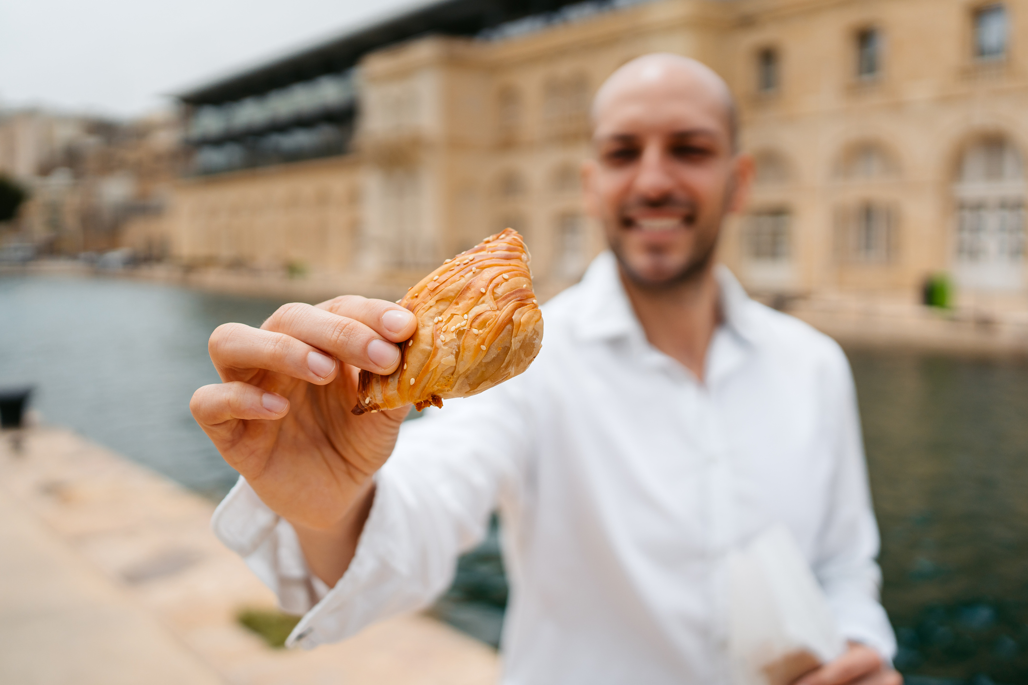 A man in a white shirt holds up a pastry by a calm waterway, smiling in front of a historic building.