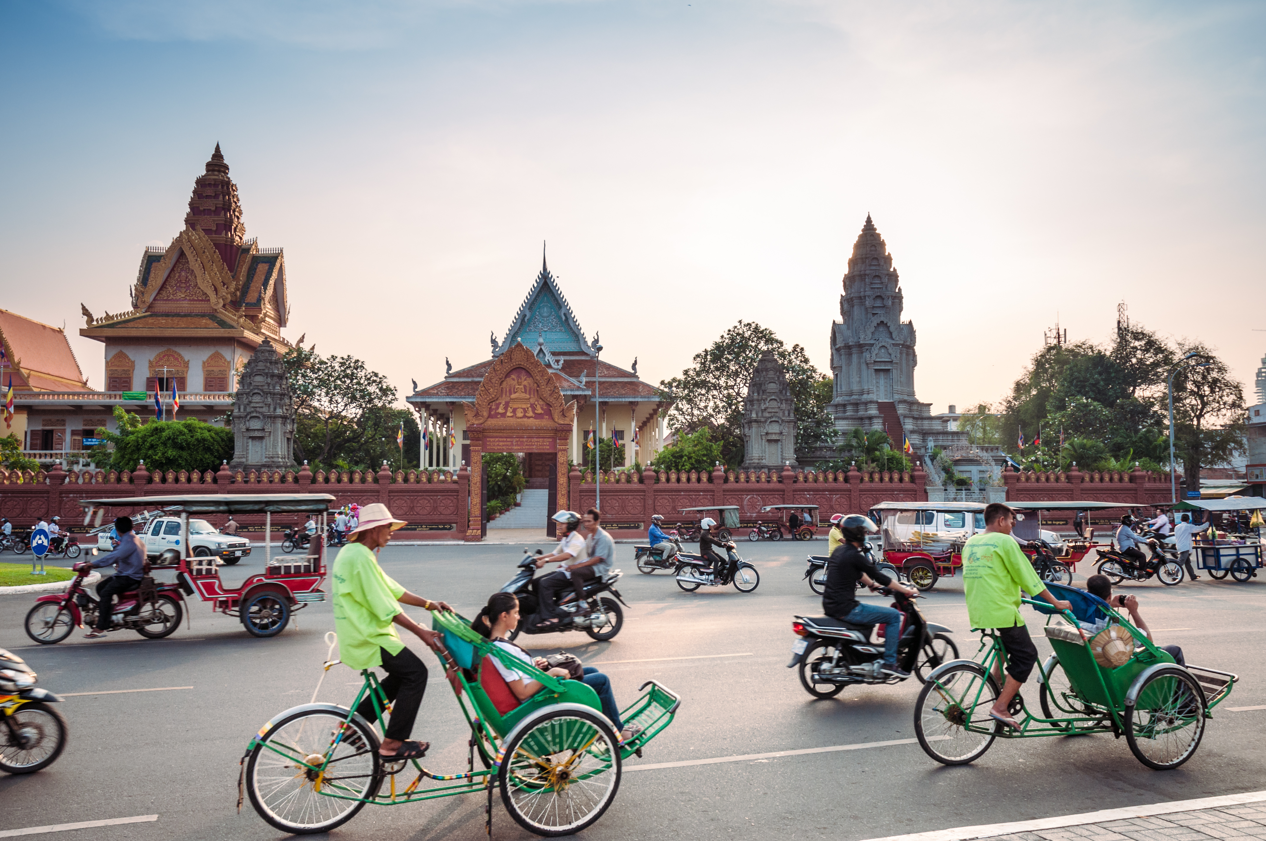 Traffic scene with rickshaws and motorbikes near a colorful temple complex at sunset.
