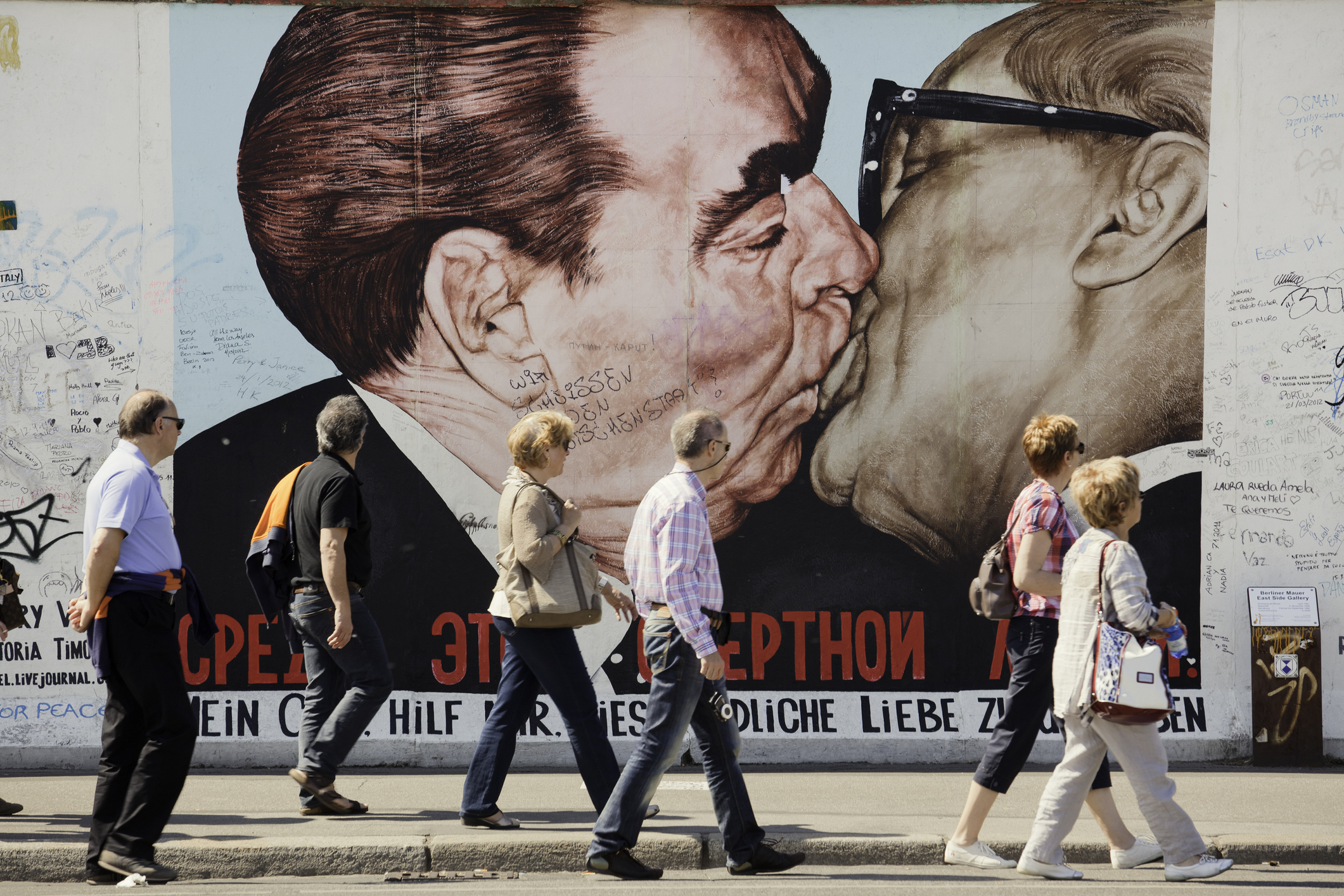 People walk by a large mural of two leaders kissing on a wall, with graffiti surrounding the artwork.