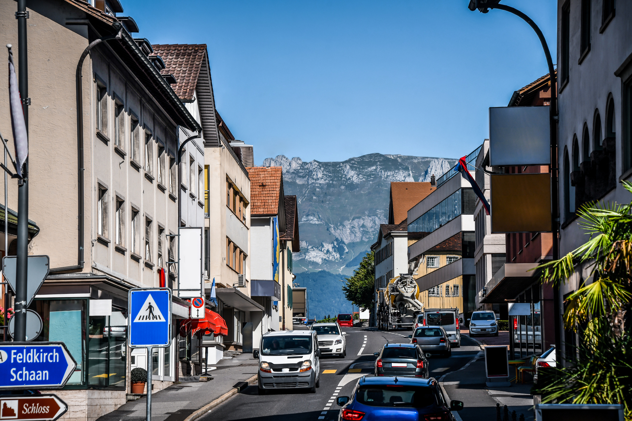 A street scene with shops, cars, and mountains in the background under a clear blue sky.