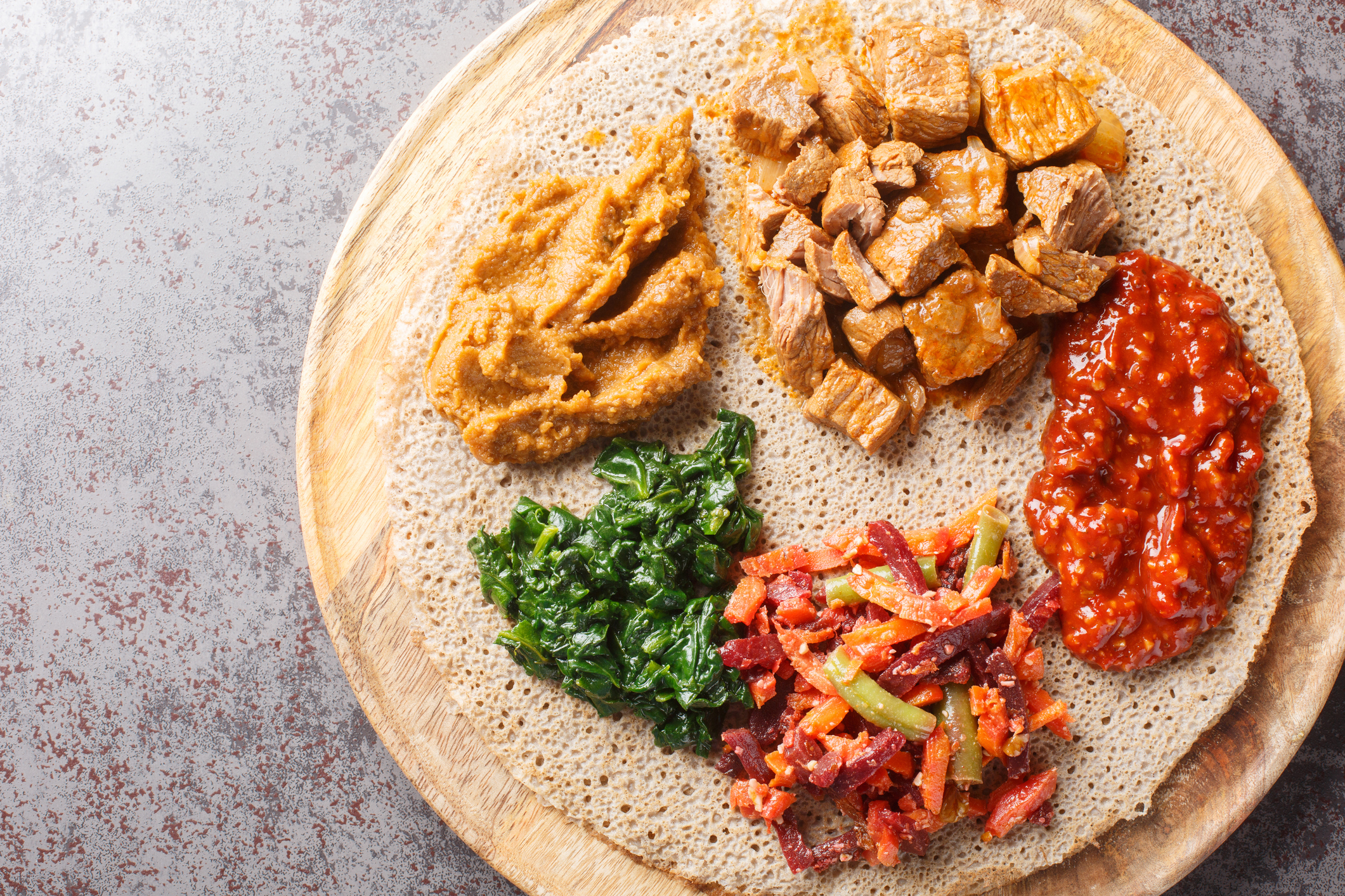 A wooden platter with injera topped with various dishes: spiced meat, lentils, greens, and a colorful vegetable mix.