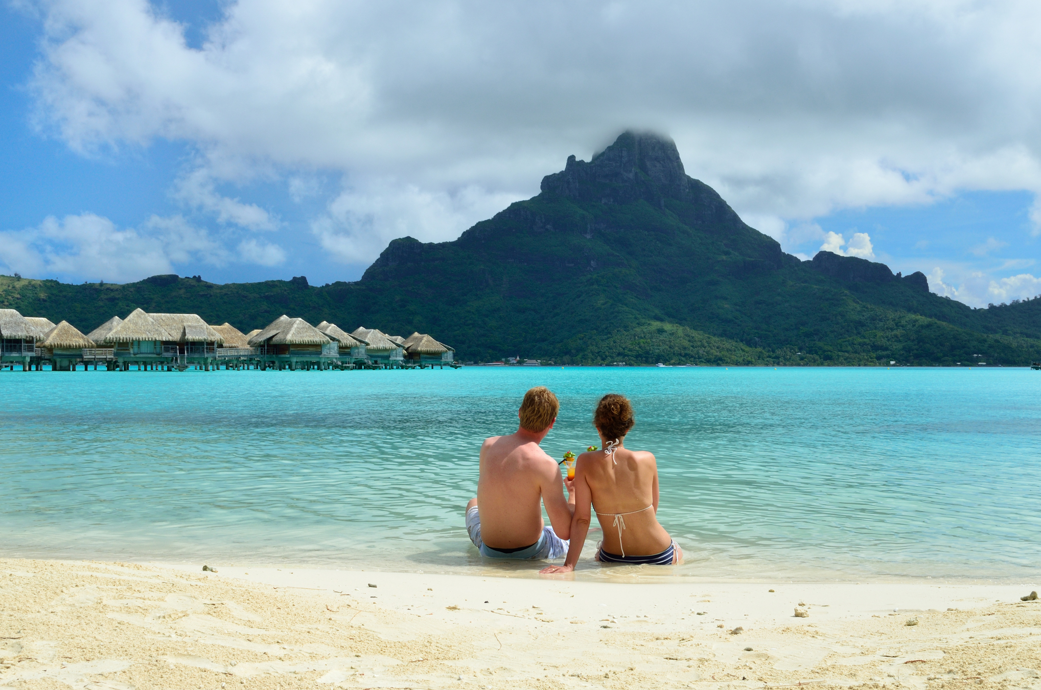 A couple sits on a beach, facing a mountain and turquoise lagoon, with huts in the background and a cloudy sky above.