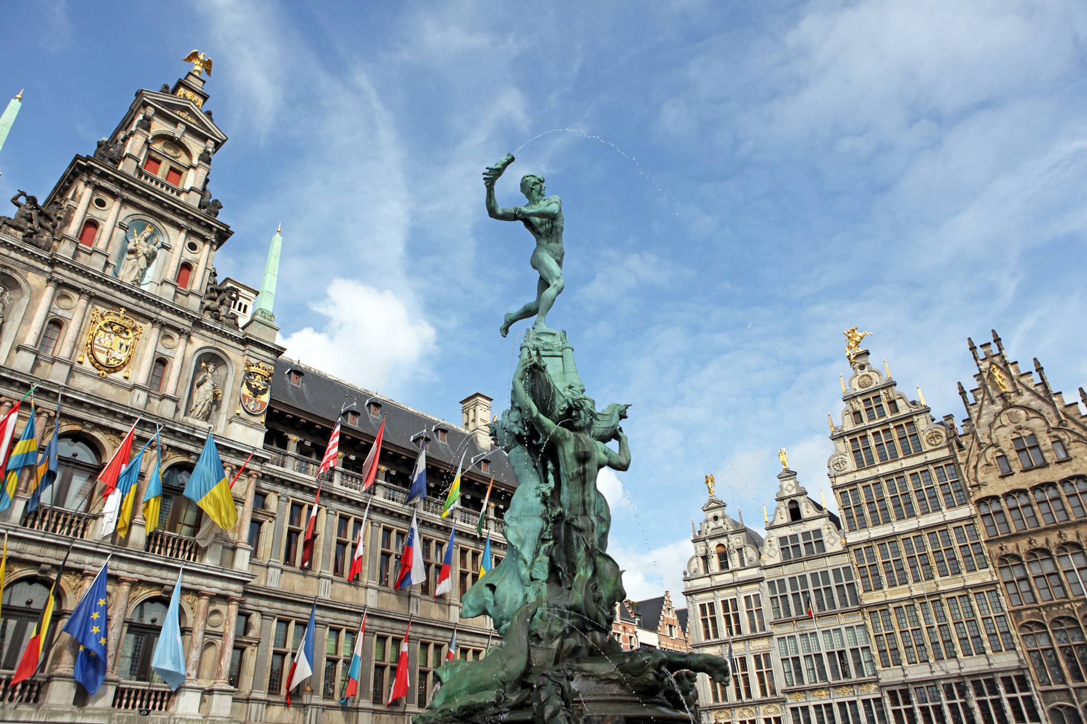 A bronze statue of a man atop a pedestal, surrounded by historic buildings and colorful flags under a blue sky.