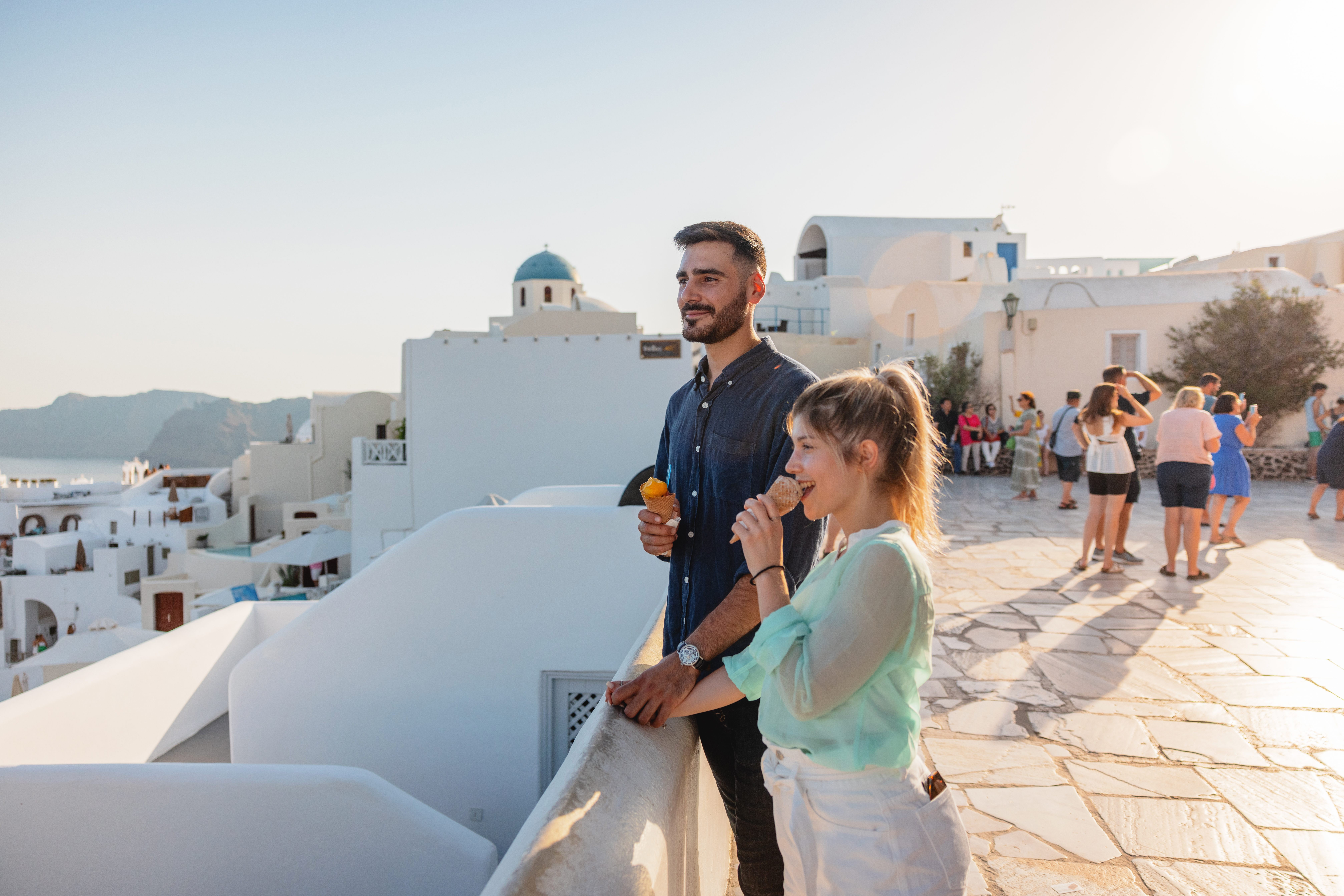 A man and a woman enjoy ice cream on a balcony, overlooking white buildings and people in a sunny coastal landscape.