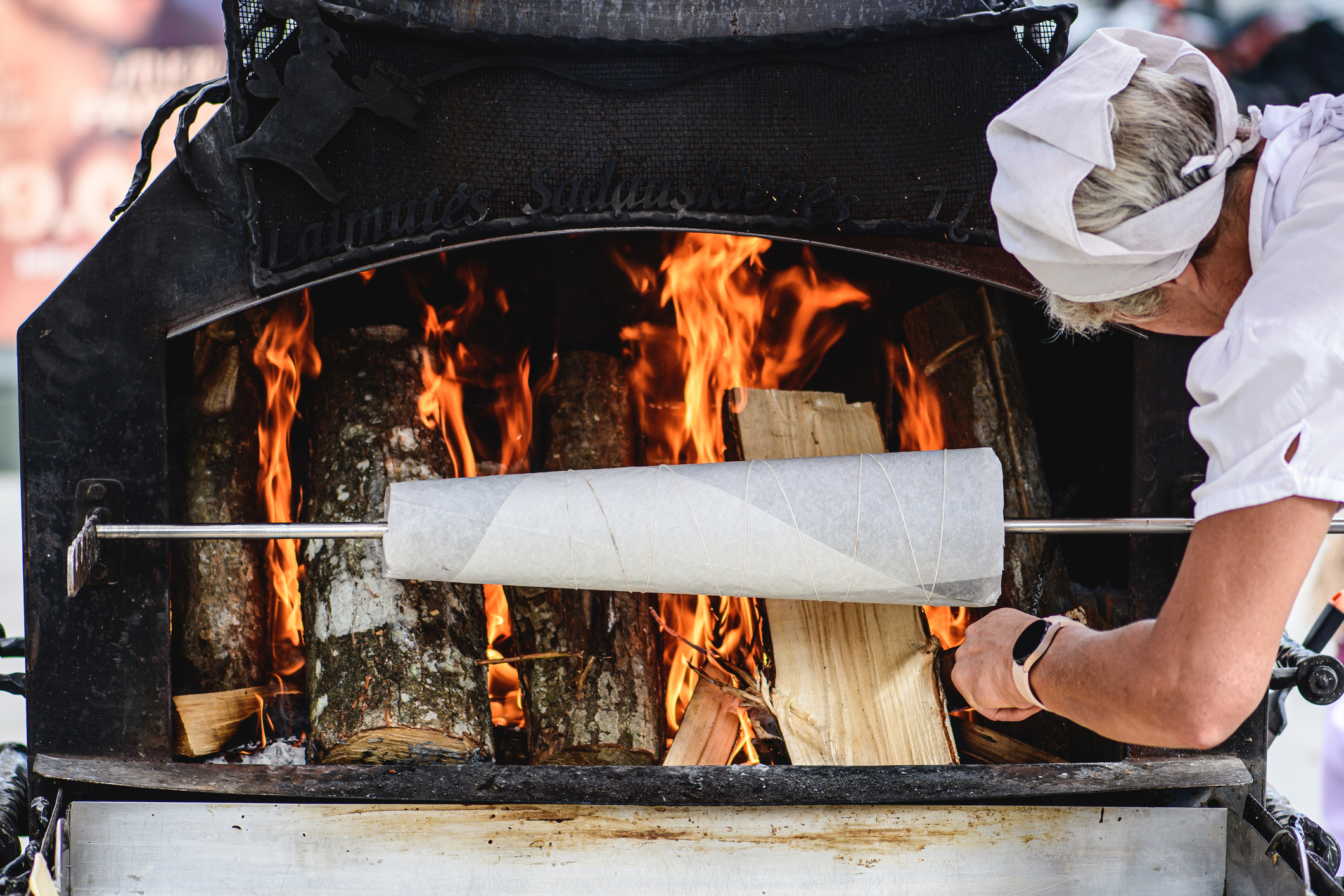 A person adjusts a cooking spit inside a wood-fired oven, with flames and logs visible in the background.