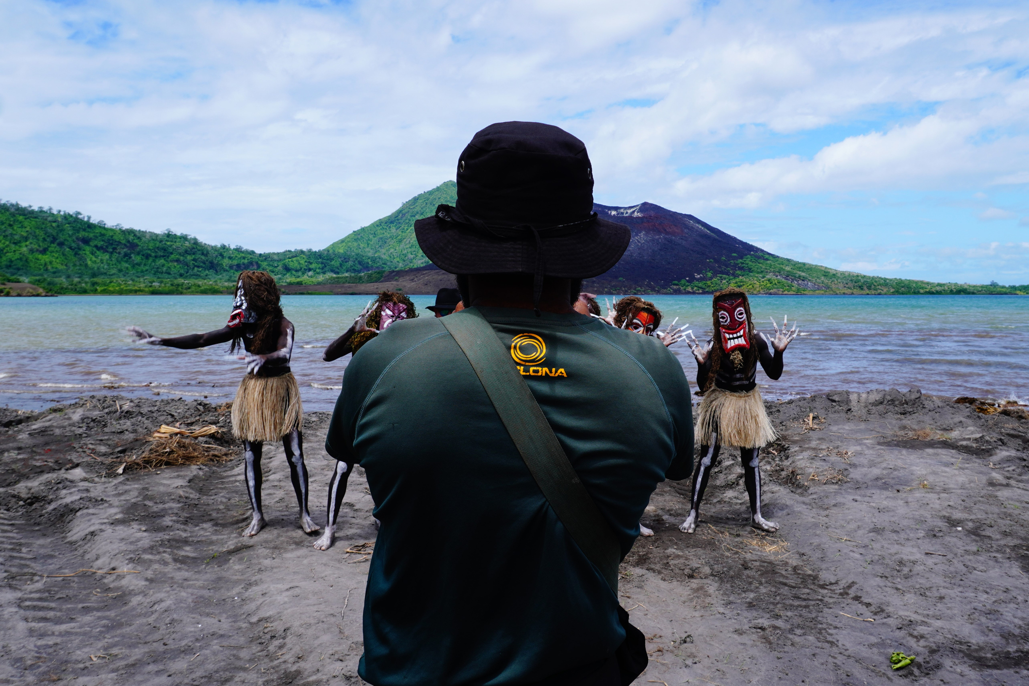 A person in a hat watches performers in masks and skirts dance by a serene waterfront and mountain backdrop.