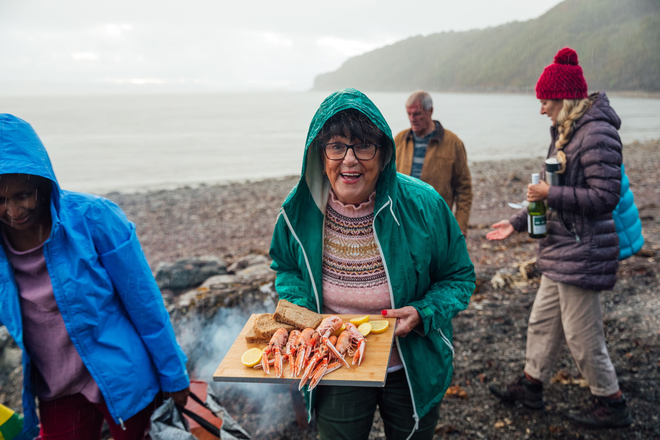 A smiling woman in a green jacket holds a tray of seafood by the beach, with friends nearby in casual attire.