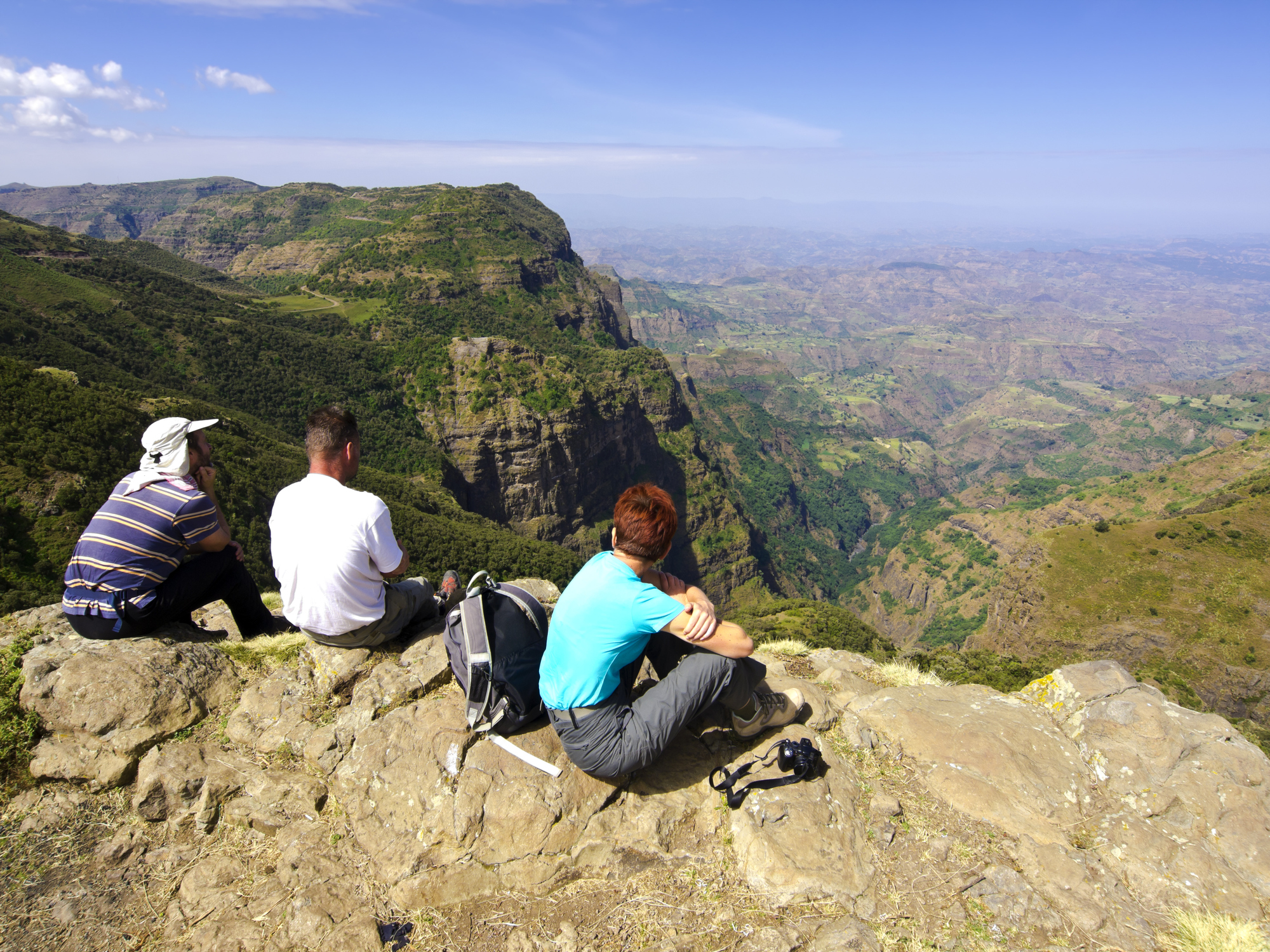 Three people sit on a rocky ledge, overlooking a vast mountainous landscape under a clear blue sky.