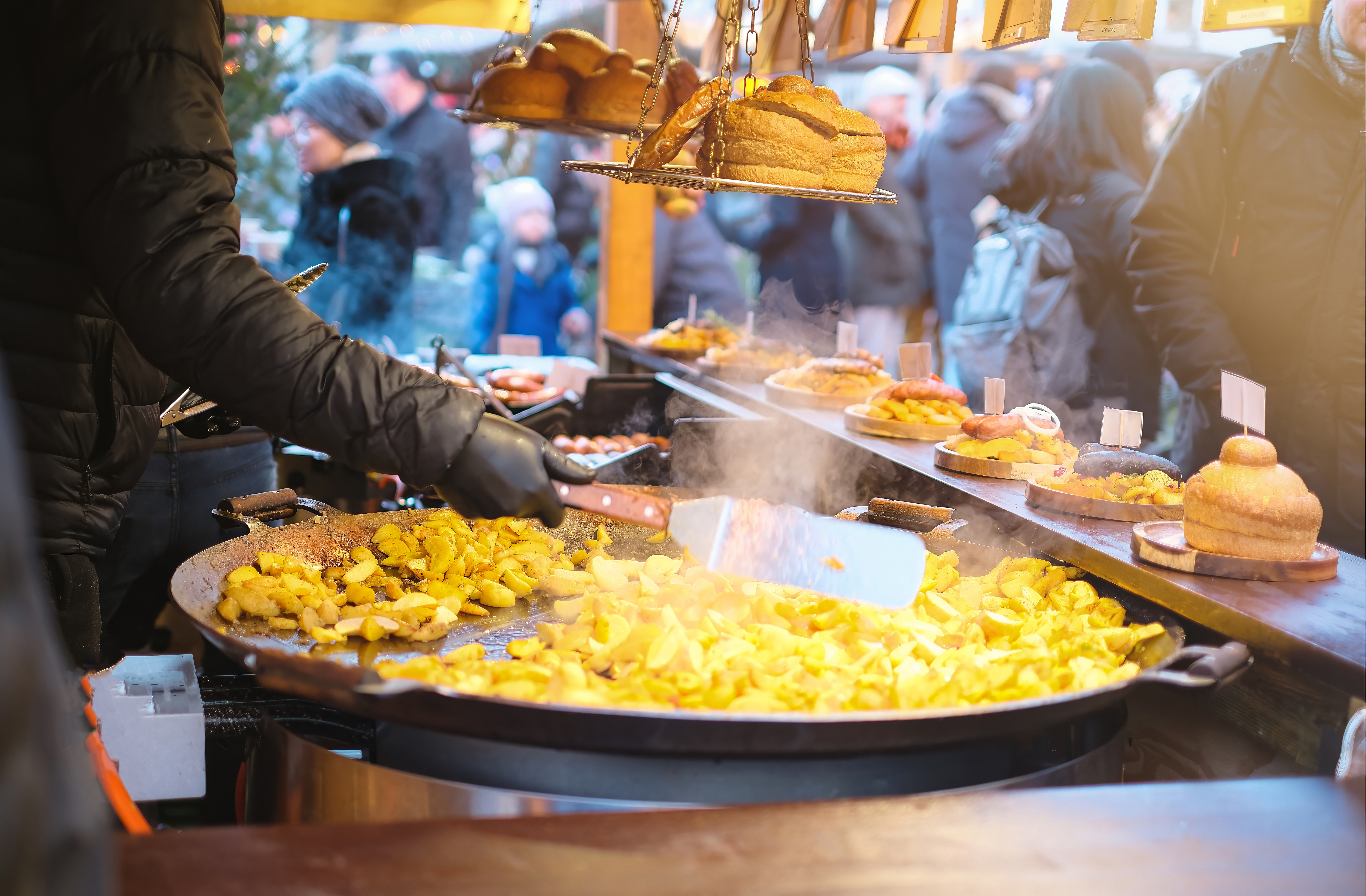 A vendor stirs a large pan of golden potatoes at a bustling outdoor market, surrounded by people and food stalls.