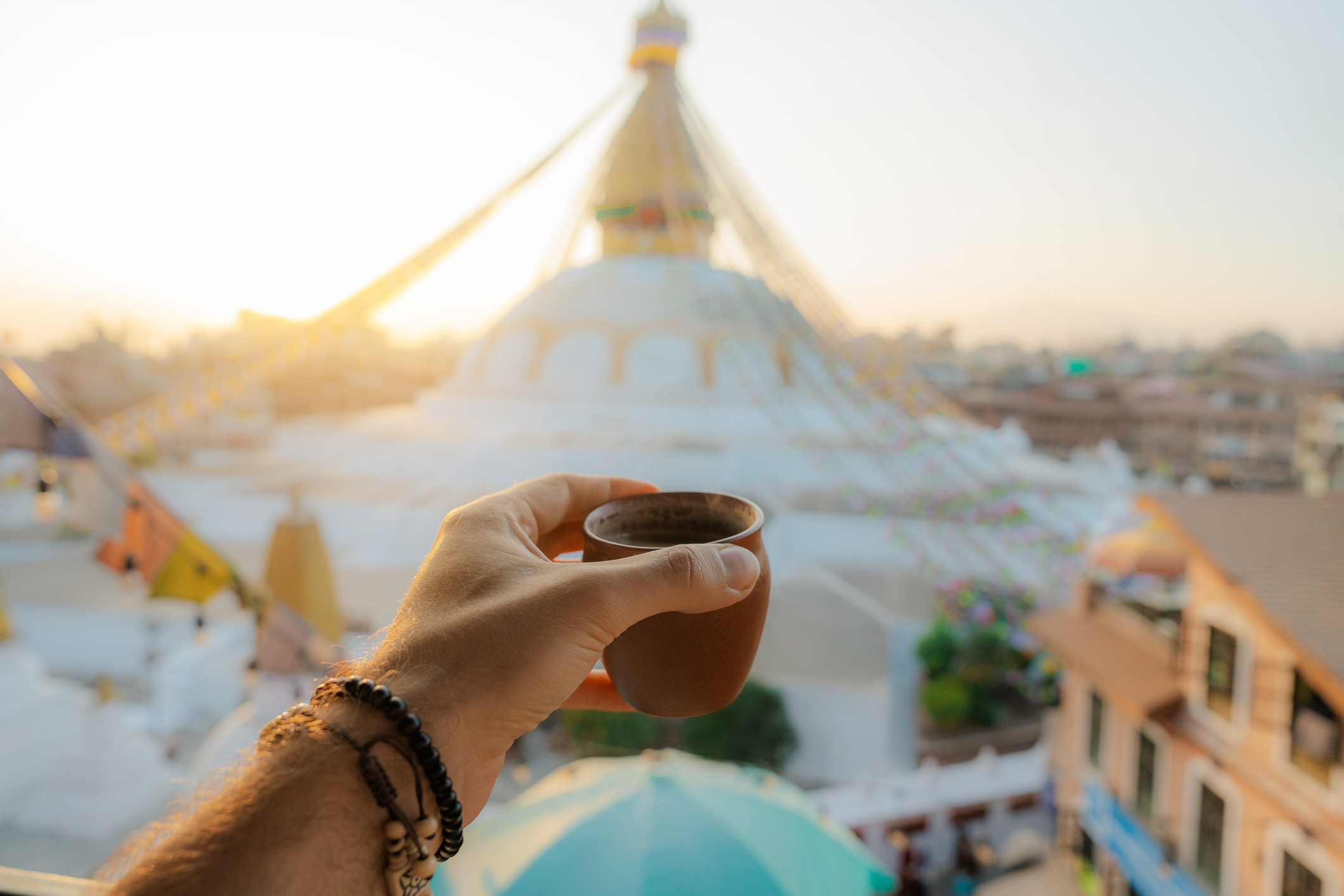 A hand holding a small clay cup in front of a stupa at sunrise, with colorful prayer flags in the background.