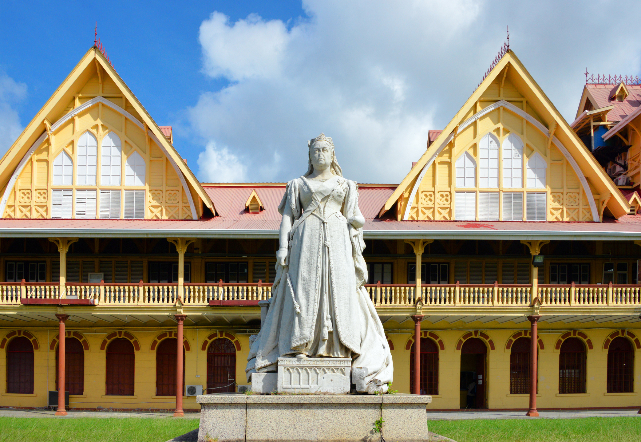 Statue of a woman in white dress in front of a historic yellow building with peaked roofs and blue sky.