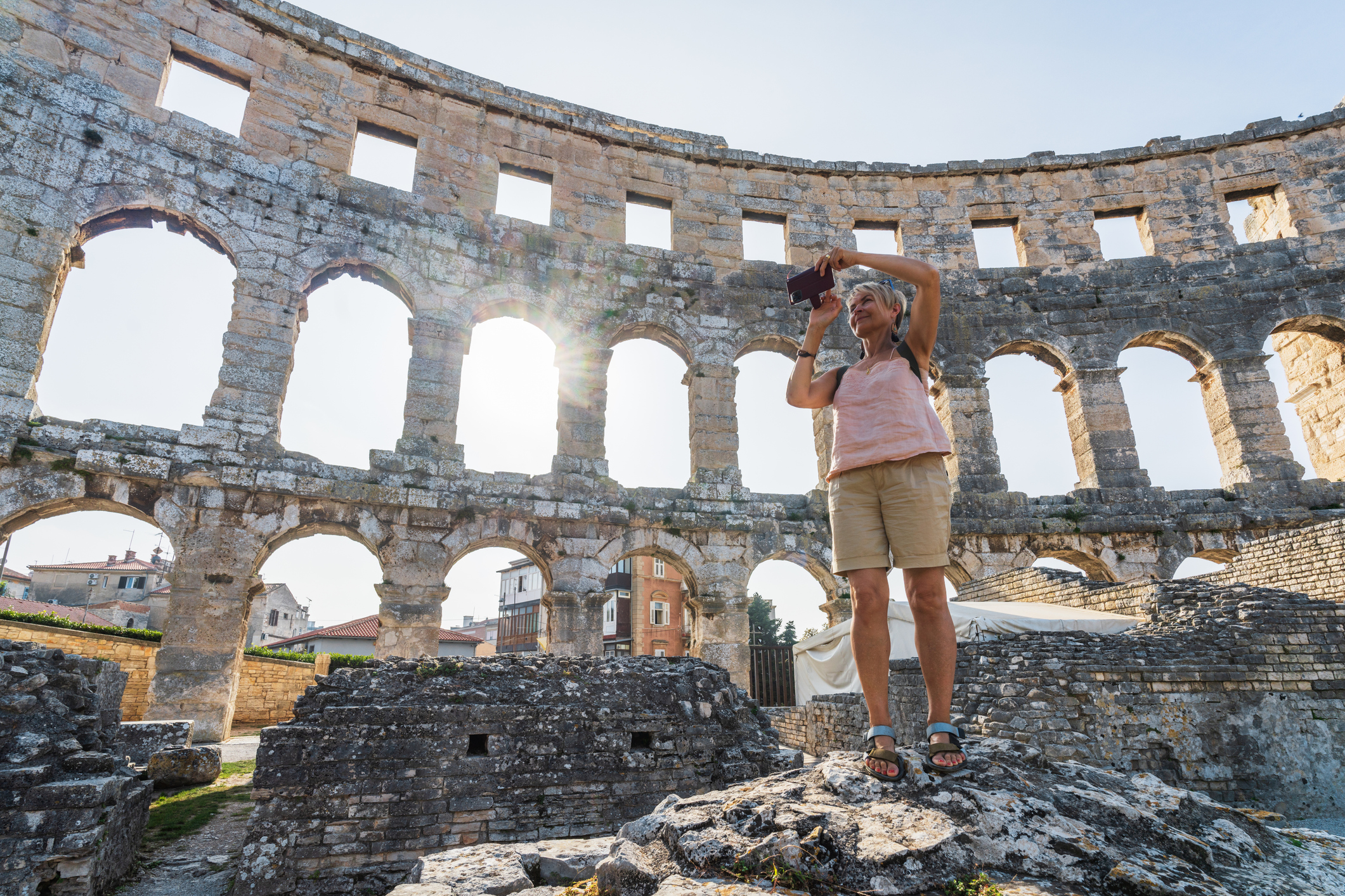 A person stands on rocks, photographing the ruins of a coliseum with sunlight streaming through the arches.