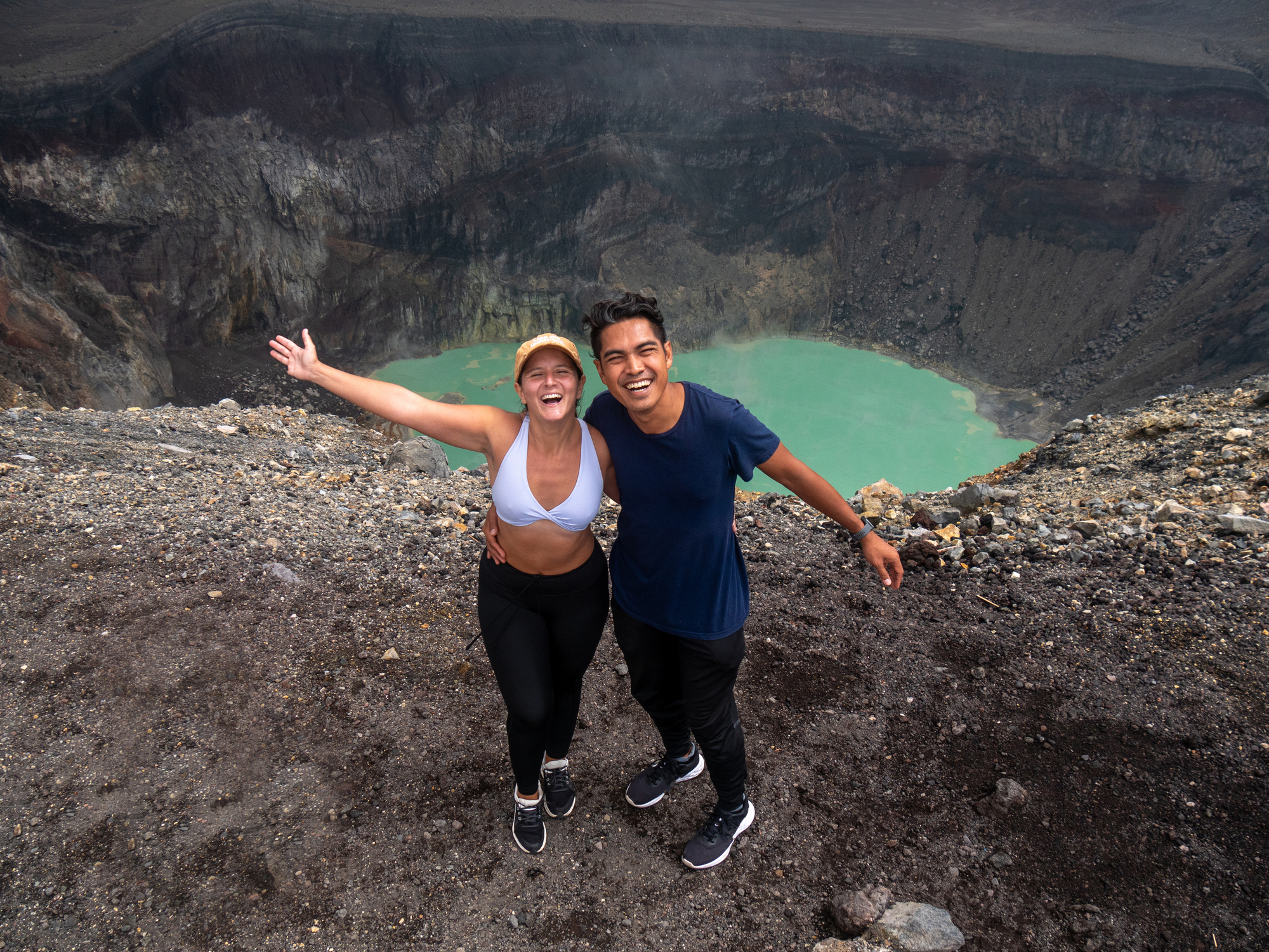 Two smiling people pose near a volcanic crater, with turquoise water visible below, against a dramatic rocky landscape.