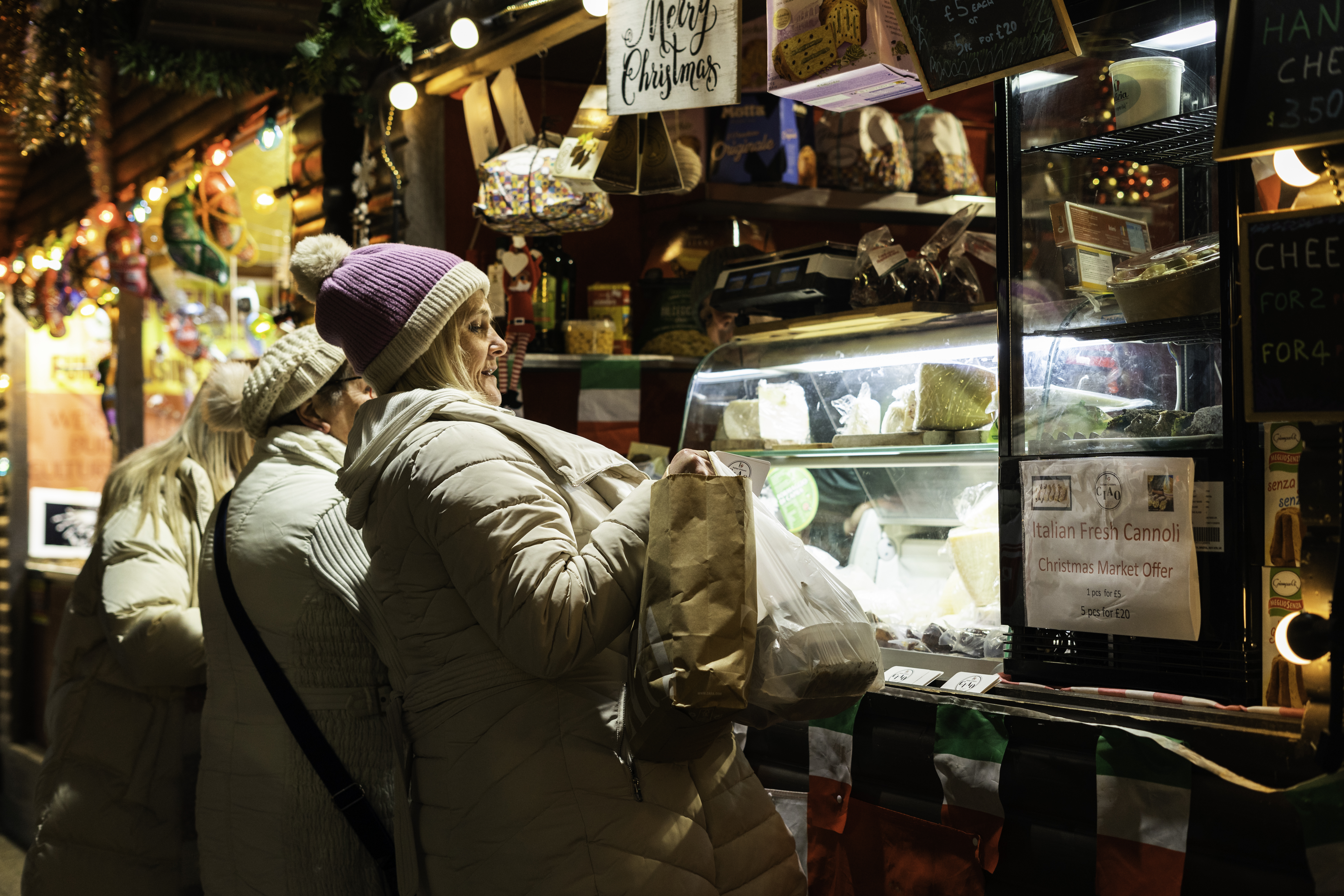 Two women in winter coats browse a festive market stall displaying various cheeses, surrounded by holiday lights.