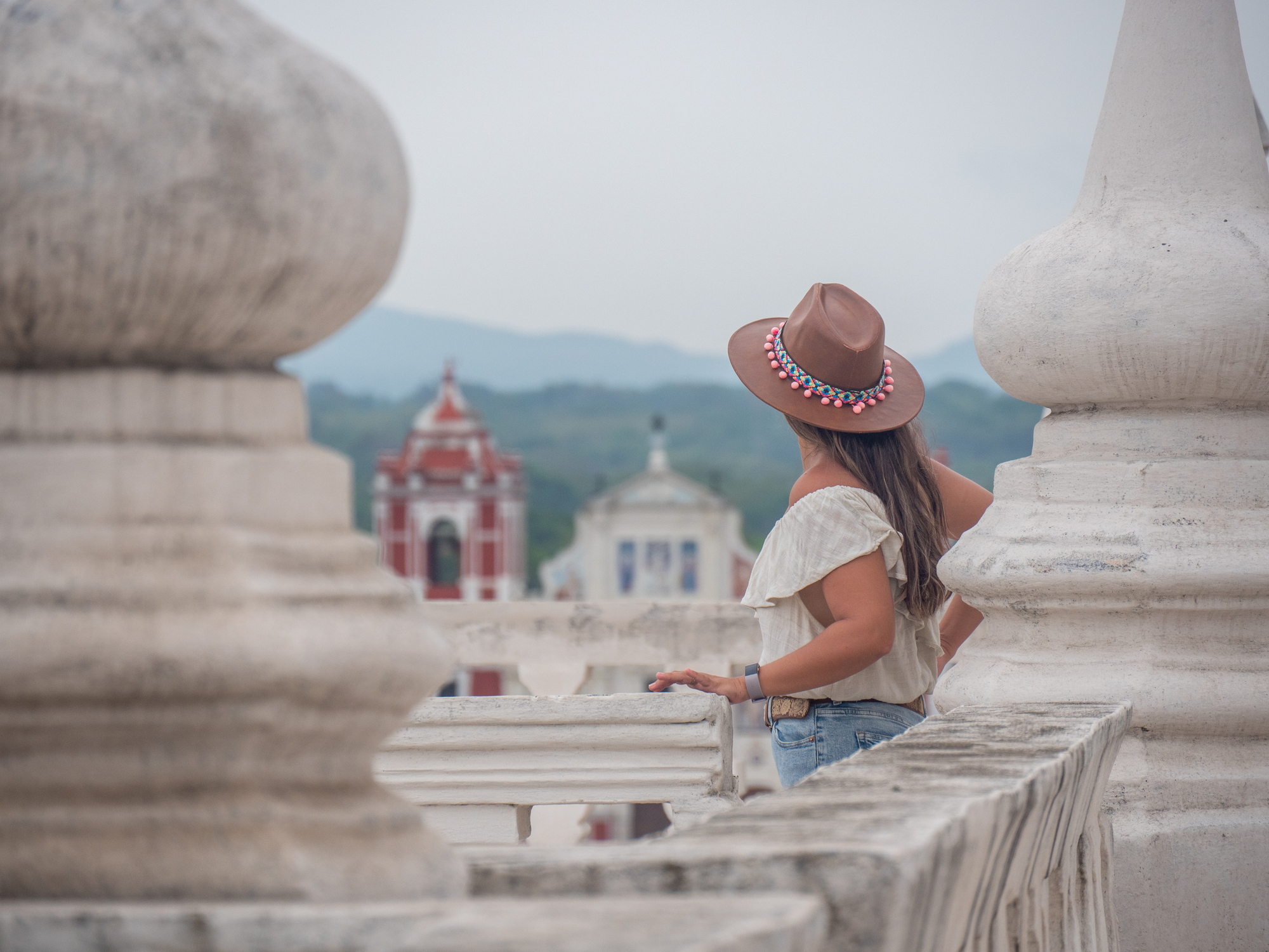A woman in a tan hat stands on a terrace, gazing at colorful buildings and mountains in the distance.